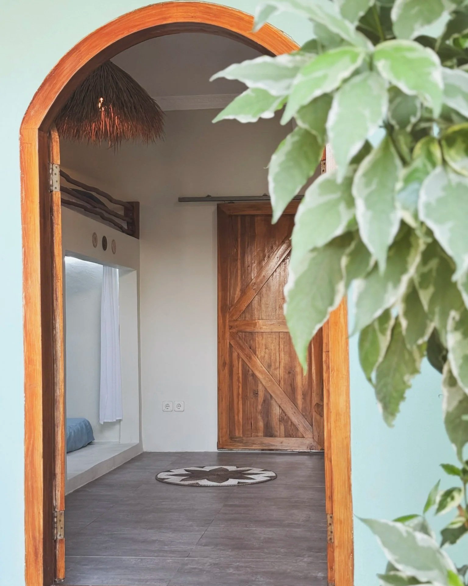 View through an arched doorway into a room with a wooden sliding door, a round patterned rug on a tiled floor, and a bed with a blue blanket in a small nook. Green leaves partially frame the right side of the image.
