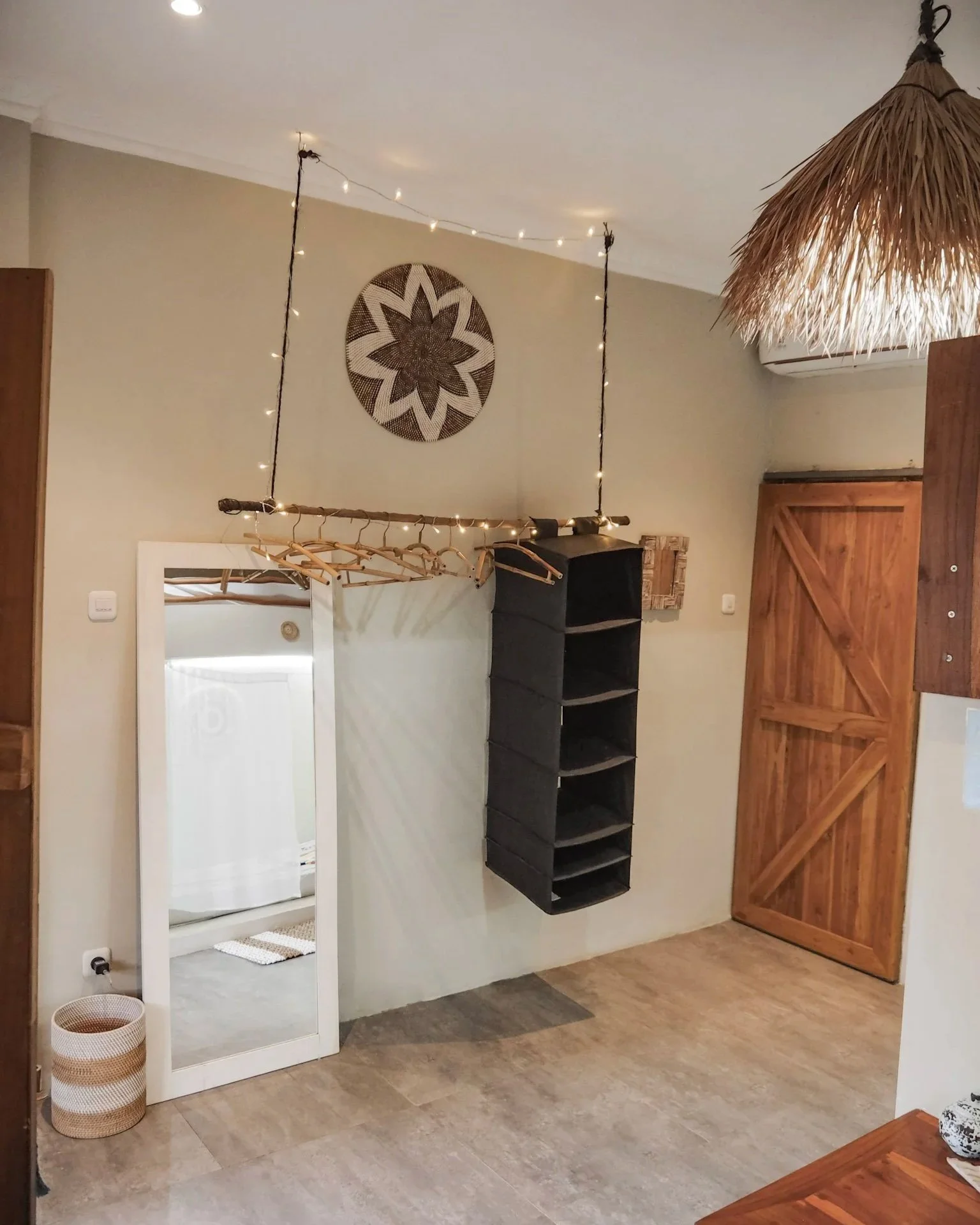 Interior of a room with a wall mirror, a hanging clothes rack with wooden hangers, a black wall-mounted shoe rack, and a rustic wooden sliding door, decorated with fairy lights and a wall hanging.