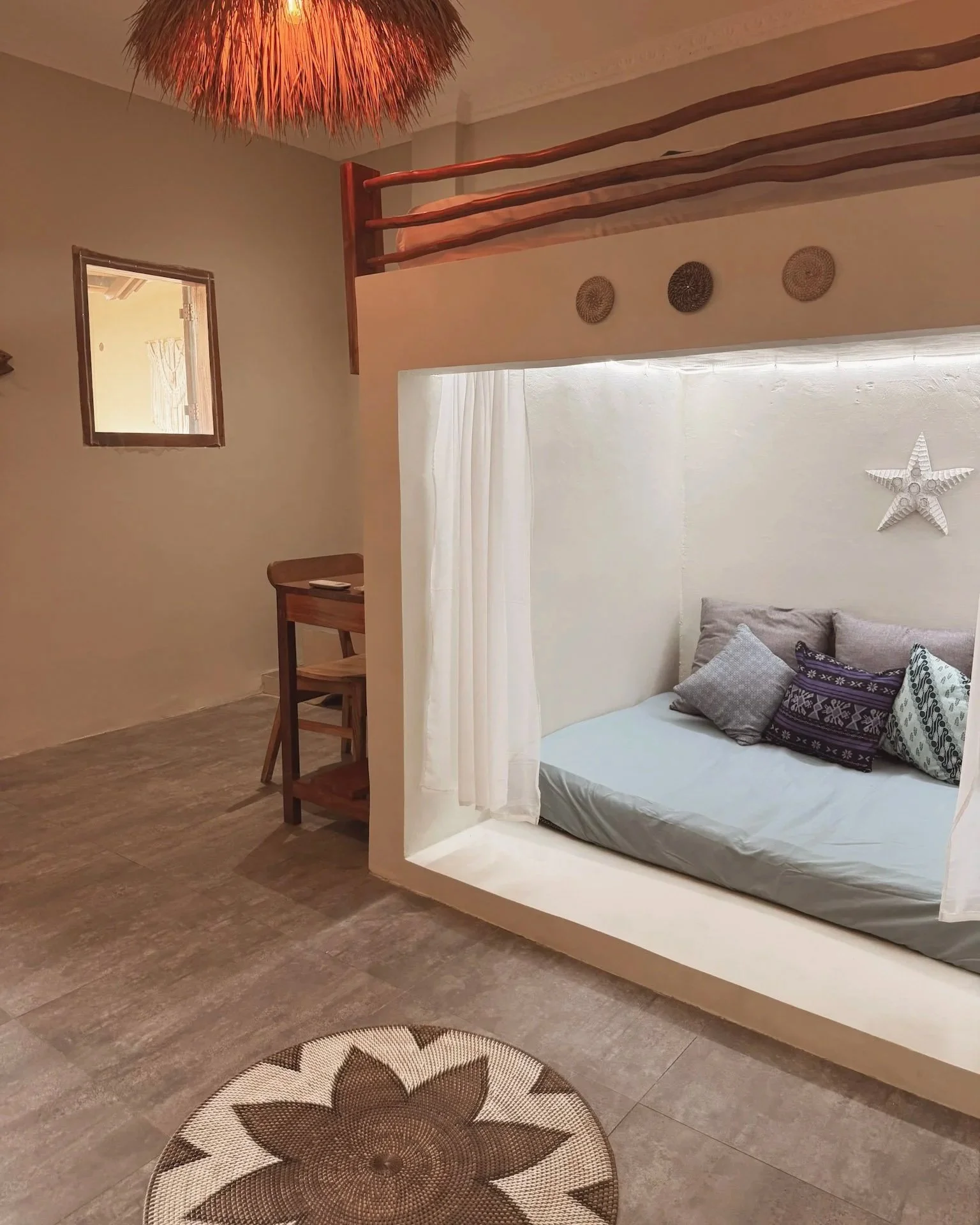 A cozy bedroom area with a built-in bed under a window and a small nook with cushions and decorative star wall art. The room has a wooden loft with a railing, a hanging lampshade made of natural fibers, tiled flooring, and a small wooden desk and cha
