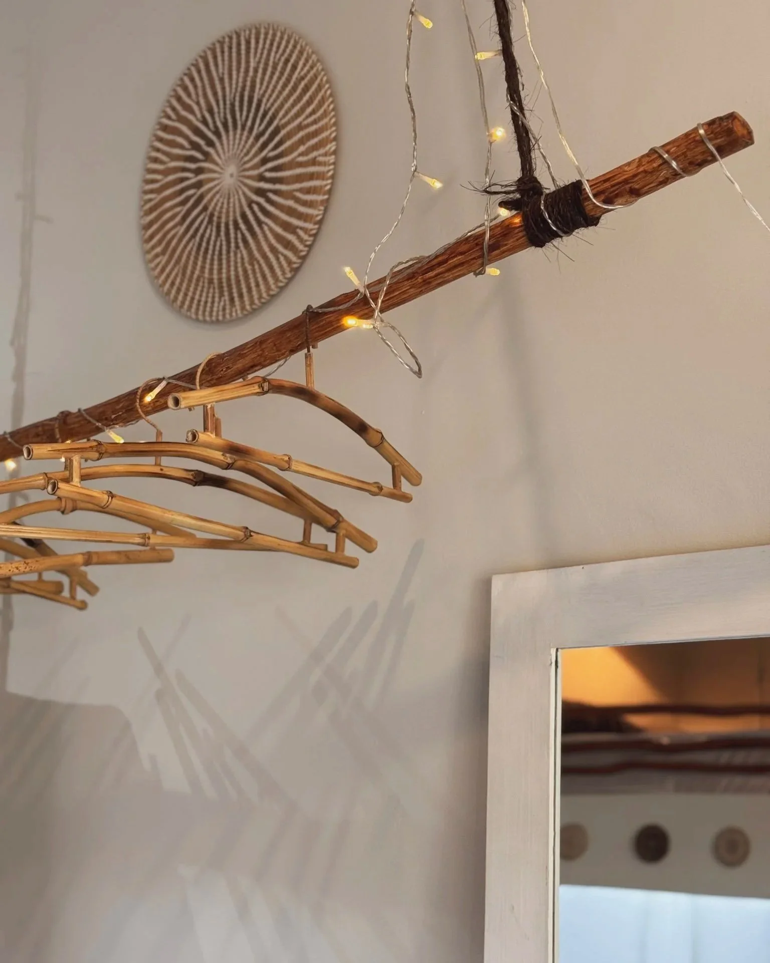 Decorative wall with a circular woven artwork, a hanging wooden and bamboo rod with string lights, and a mirror reflecting part of the room.