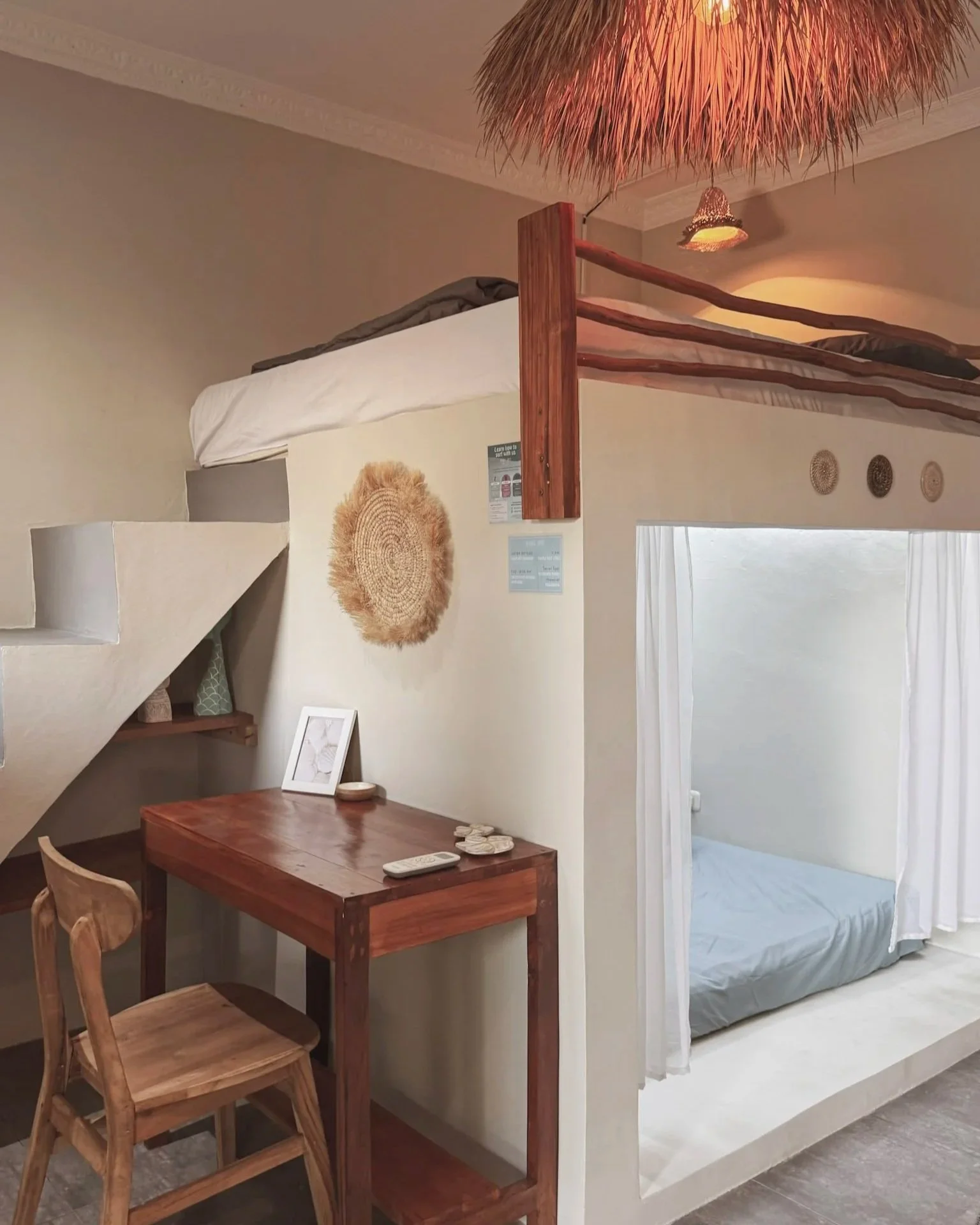 A cozy hotel room with a loft bed accessible by stairs, a small wooden desk with a chair, and a separate bed area with a curtain divider, decorated in natural and neutral tones.