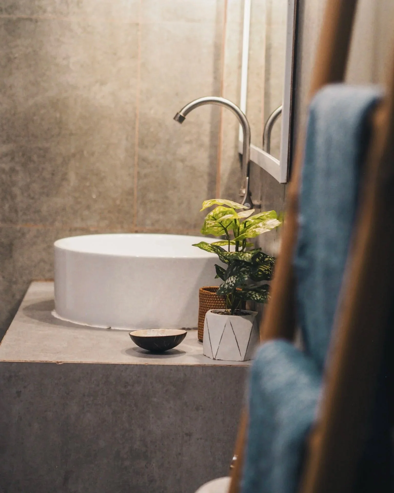 Modern bathroom sink with a white vessel basin, a silver faucet, a potted plant, a decorative bowl, and a mirror in the background.