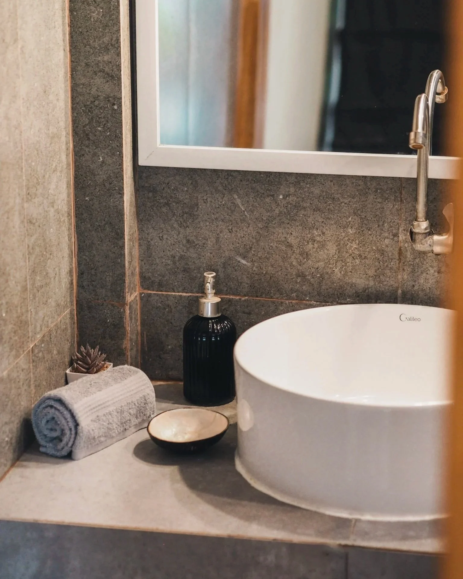 Bathroom sink area with a white round vessel sink, gray countertop, black soap dispenser, rolled gray towel, small plant, and decorative bowl, reflected in mirror.