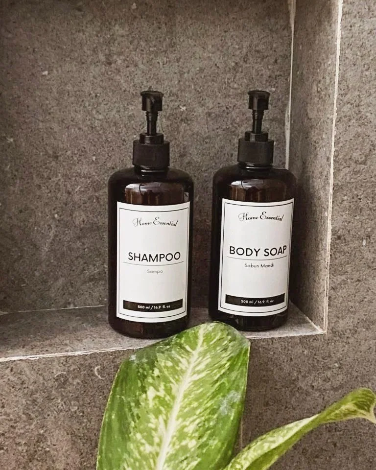 Two brown bottles with pumps labeled 'Shampoo' and 'Body Soap' on a tiled shower wall, with a green and white plant at the bottom of the image.