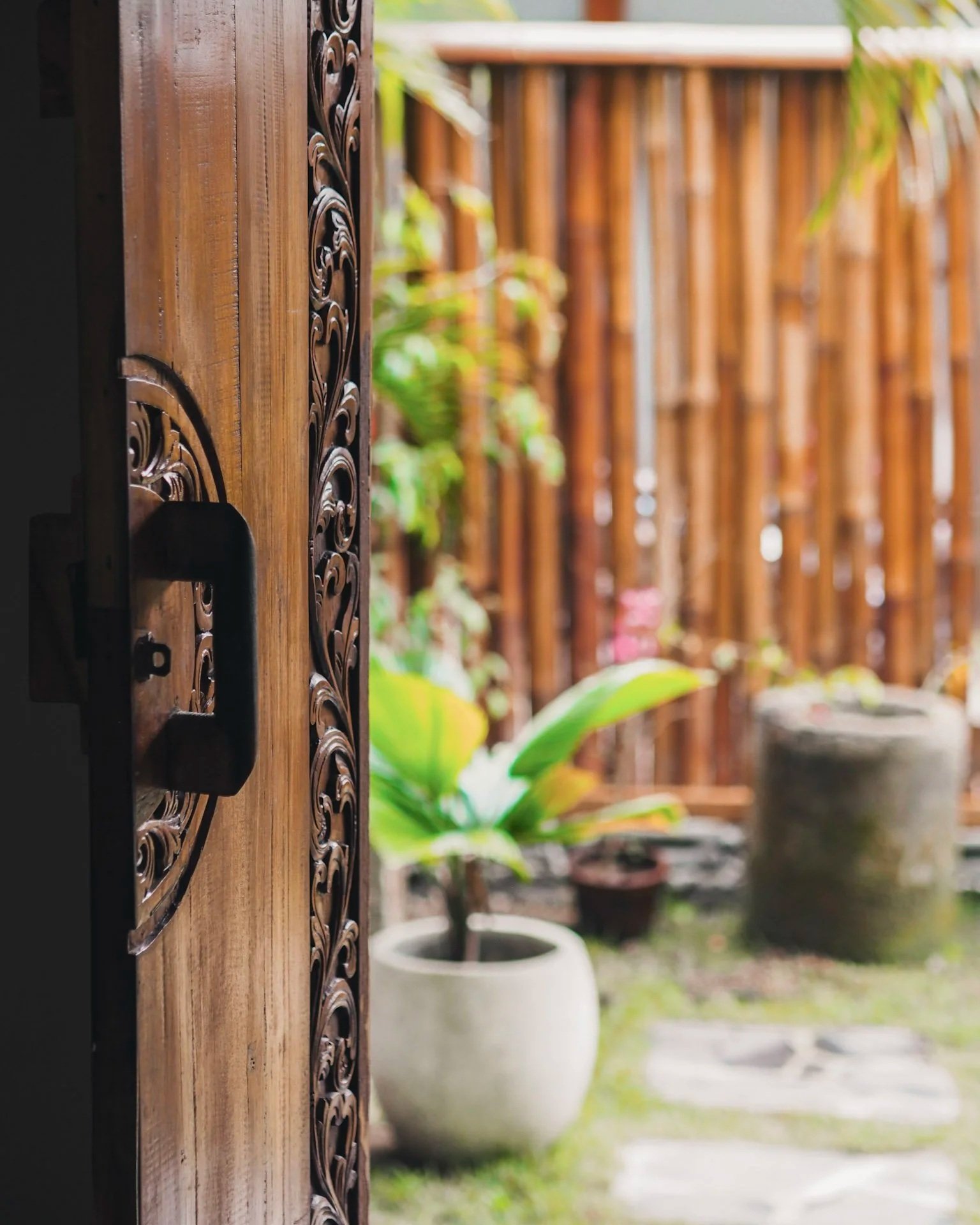 View of a garden through an open wooden door with intricate carvings, showing potted plants and bamboo fencing in the background.