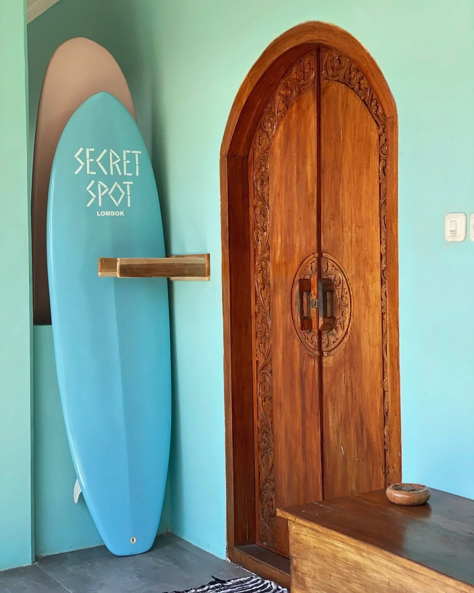 A blue surfboard with the words 'Secret Spot Lombok' written on it, leaning against a light blue wall beside an ornate wooden door with intricate carvings.