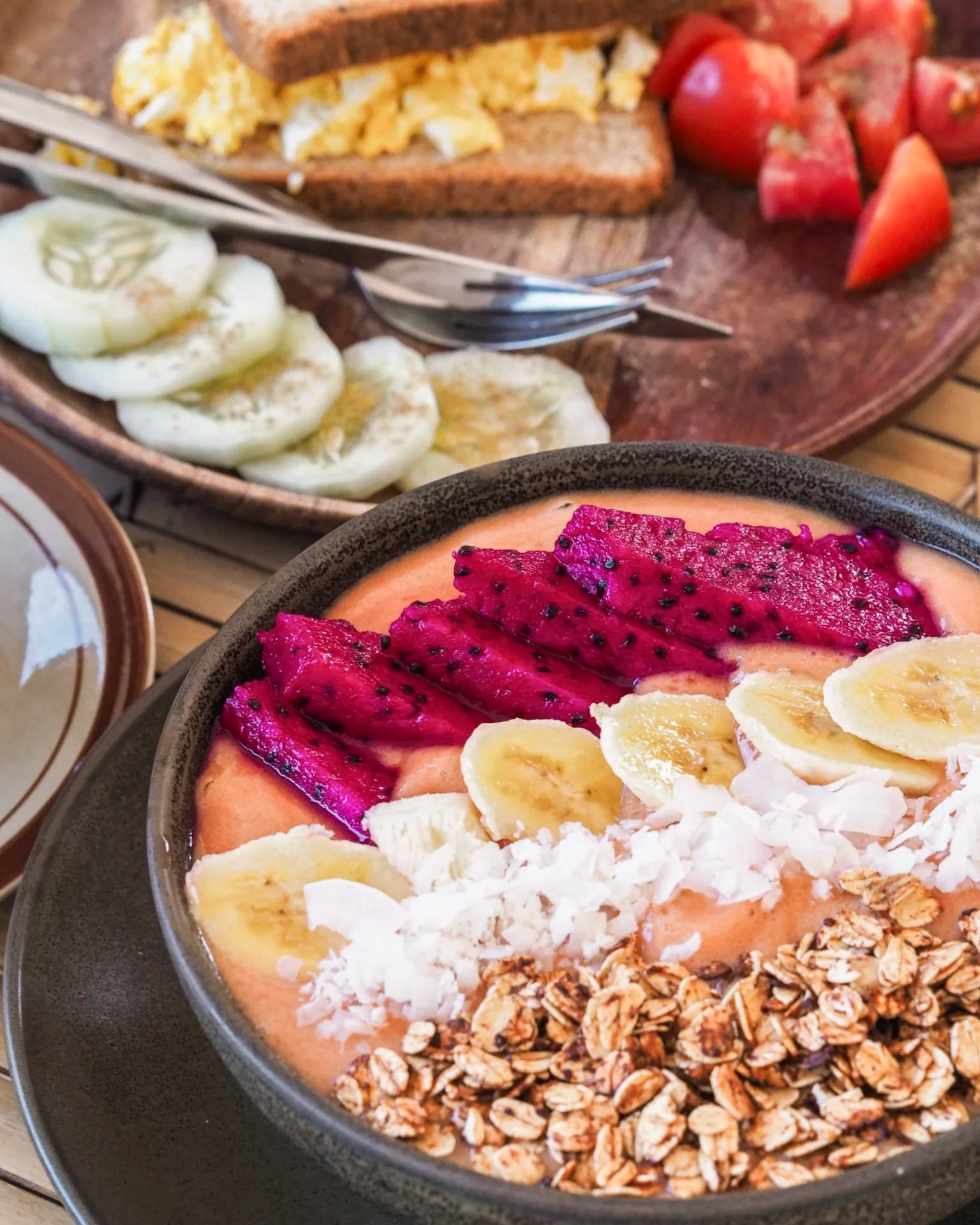 A bowl of smoothie topped with sliced bananas, dragon fruit, shredded coconut, and granola, placed on a tray. In the background, there is a wooden board with sliced cucumbers, scrambled eggs on toast, and sliced tomatoes with forks.