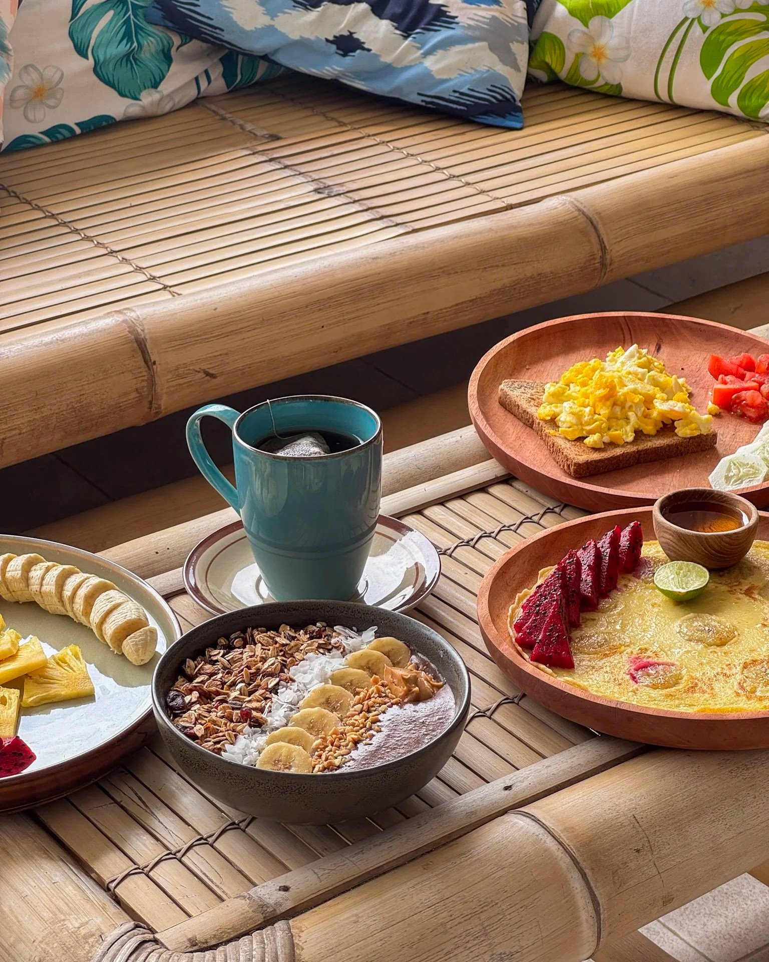 A breakfast spread on a bamboo table includes a bowl of yogurt with sliced bananas, granola, coconut, and nuts; a plate of sliced bananas and pineapple; a teacup of tea with a tea bag; a wooden bowl of scrambled eggs with toast and chopped tomatoes; 