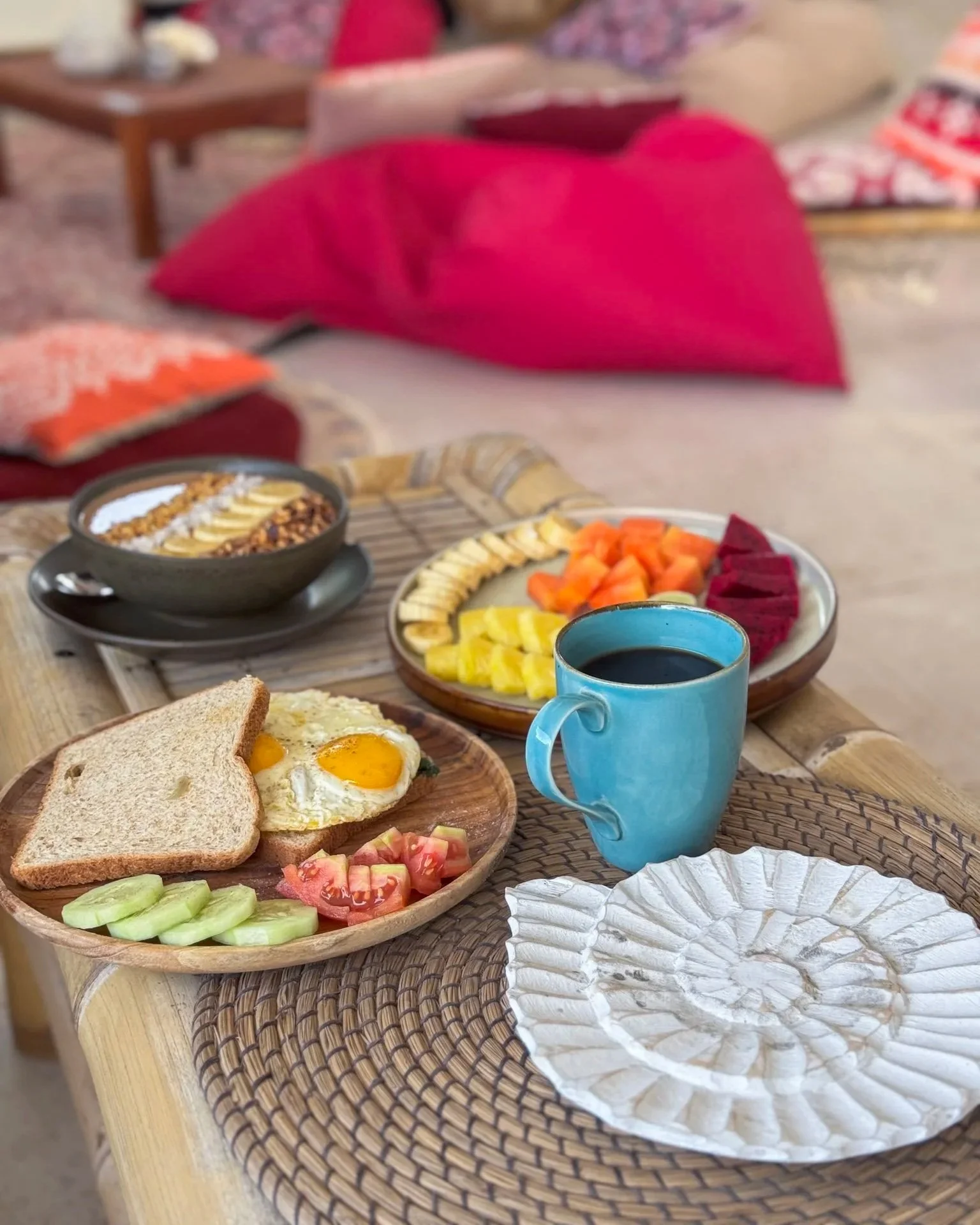 Breakfast spread on a wooden table includes fried eggs, toast, sliced cucumbers, tomatoes, and various fruits on a plate behind, a bowl of cereal and fruit, a cup of black coffee in a blue mug, and a paper plate on a woven placemat, with colorful cus