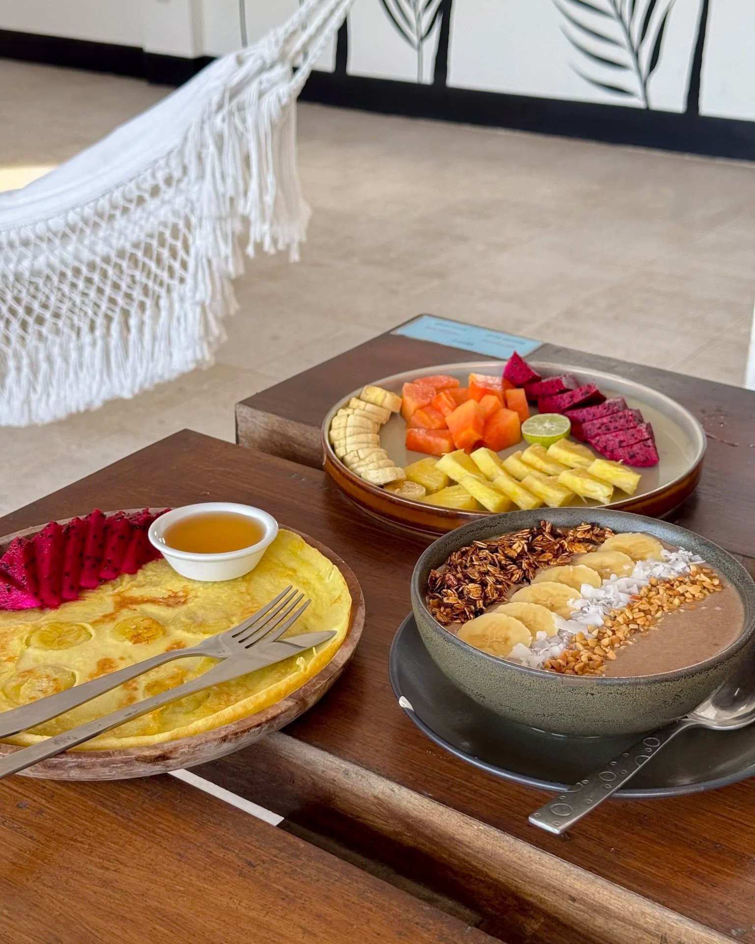 A wooden table with a bowl of smoothie topped with granola, banana, and coconut flakes, a fruit platter with sliced dragon fruit, watermelon, pineapple, honeydew, banana, and a lime, and a plate of pineapple slices with a small dish of honey.