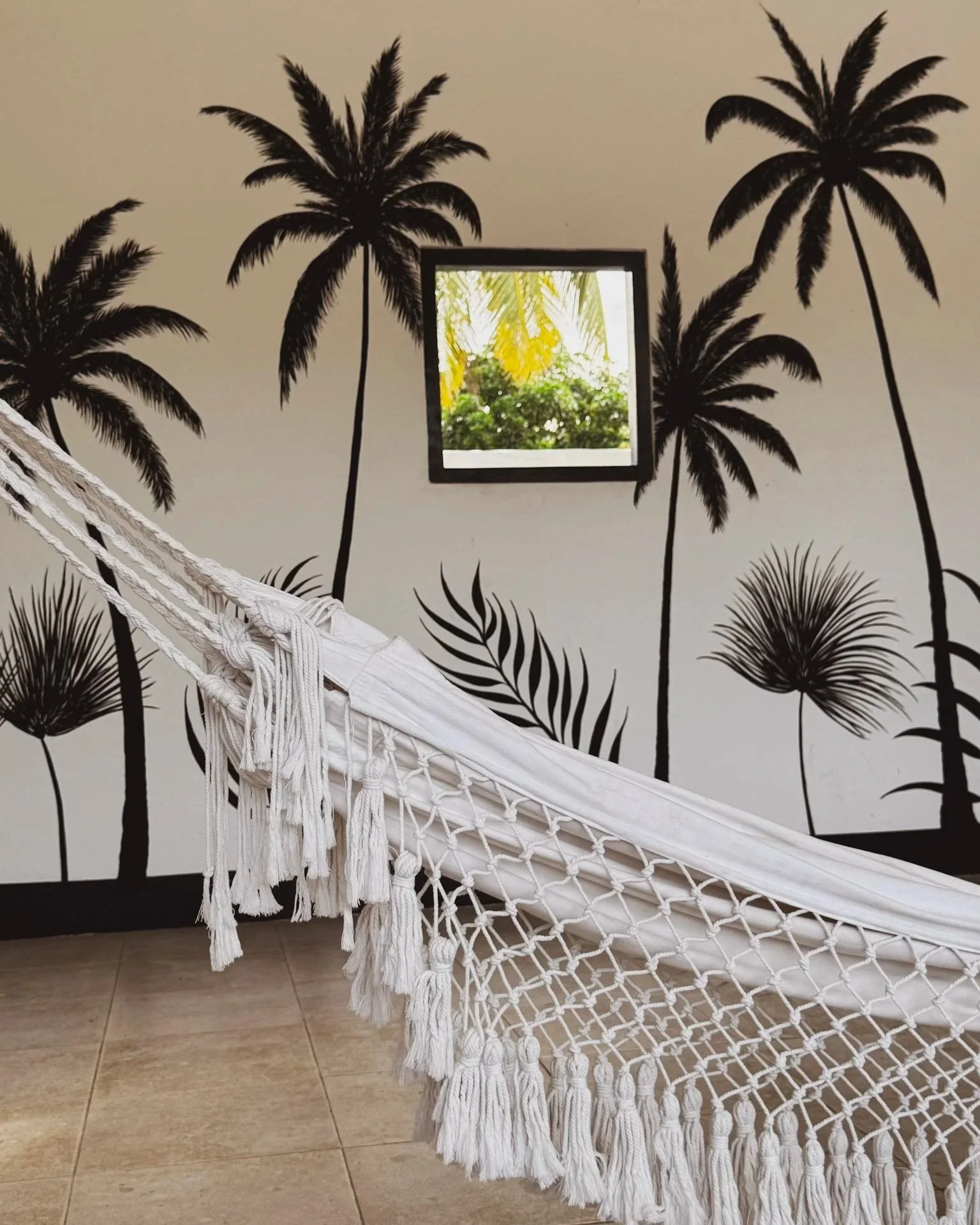 White hammock with tassels in front of a black and white tropical palm tree wall mural with a small window showing green foliage outside.