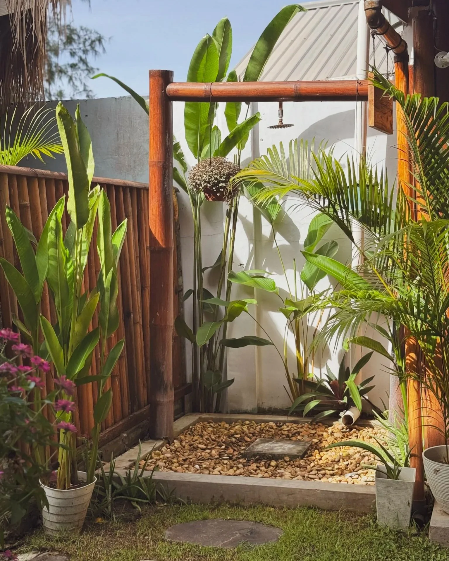 Outdoor shower with a wooden frame, surrounded by potted plants and greenery, on a small gravel yard with stepping stones.