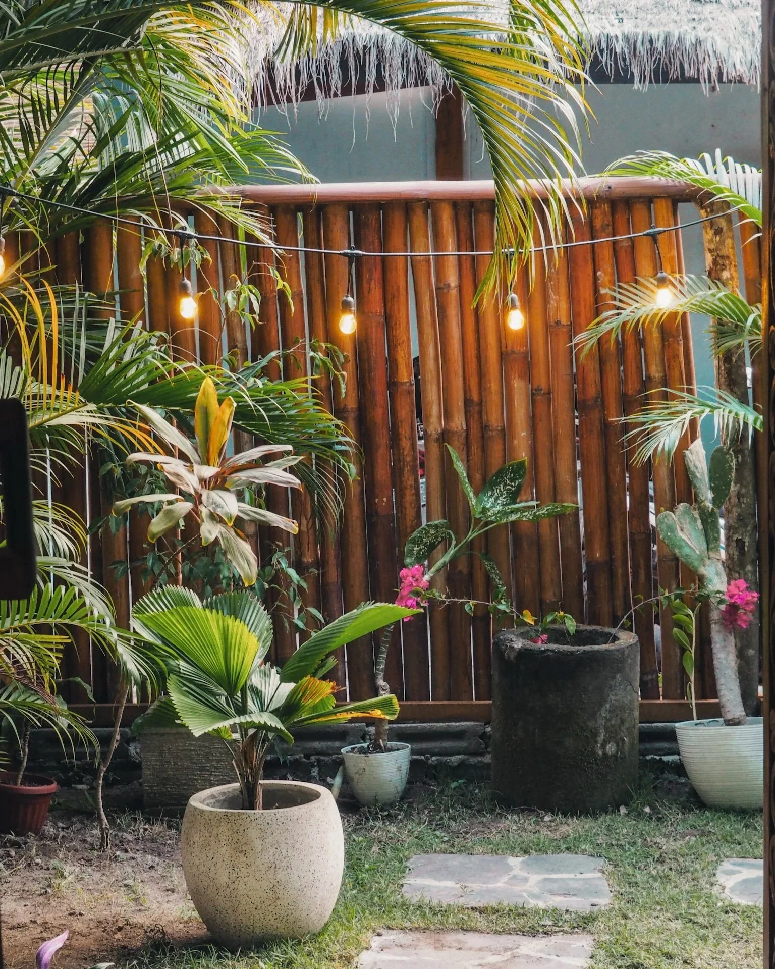 A cozy outdoor garden area with potted tropical plants and flowers, a wooden fence, string lights, and a stone pathway.