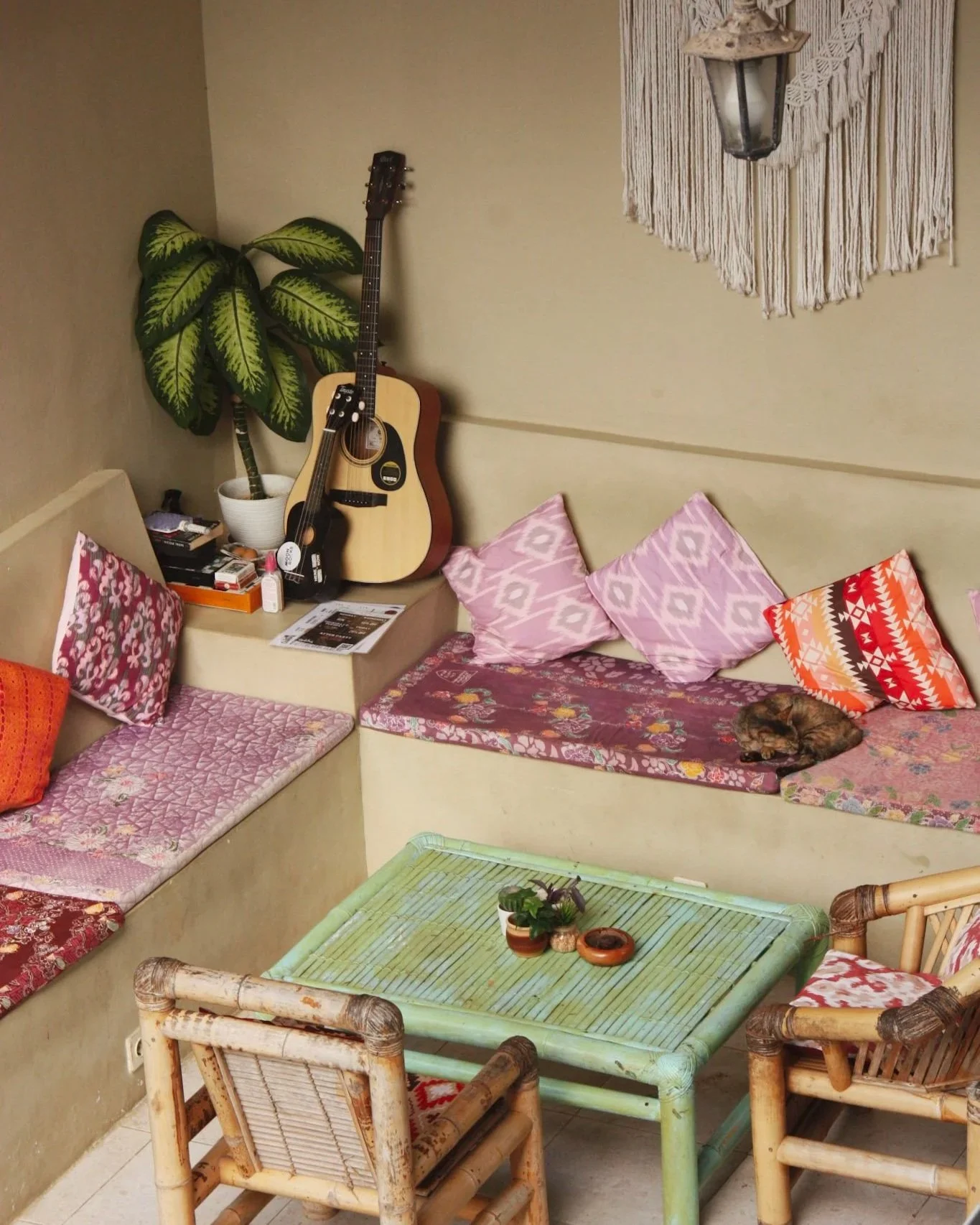 A cozy indoor corner with a built-in cushioned seating area decorated with patterned pillows, a small green coffee table with plants, a guitar and a potted plant in the corner, and a sleeping cat on the cushions. There's a wall decoration with a hang