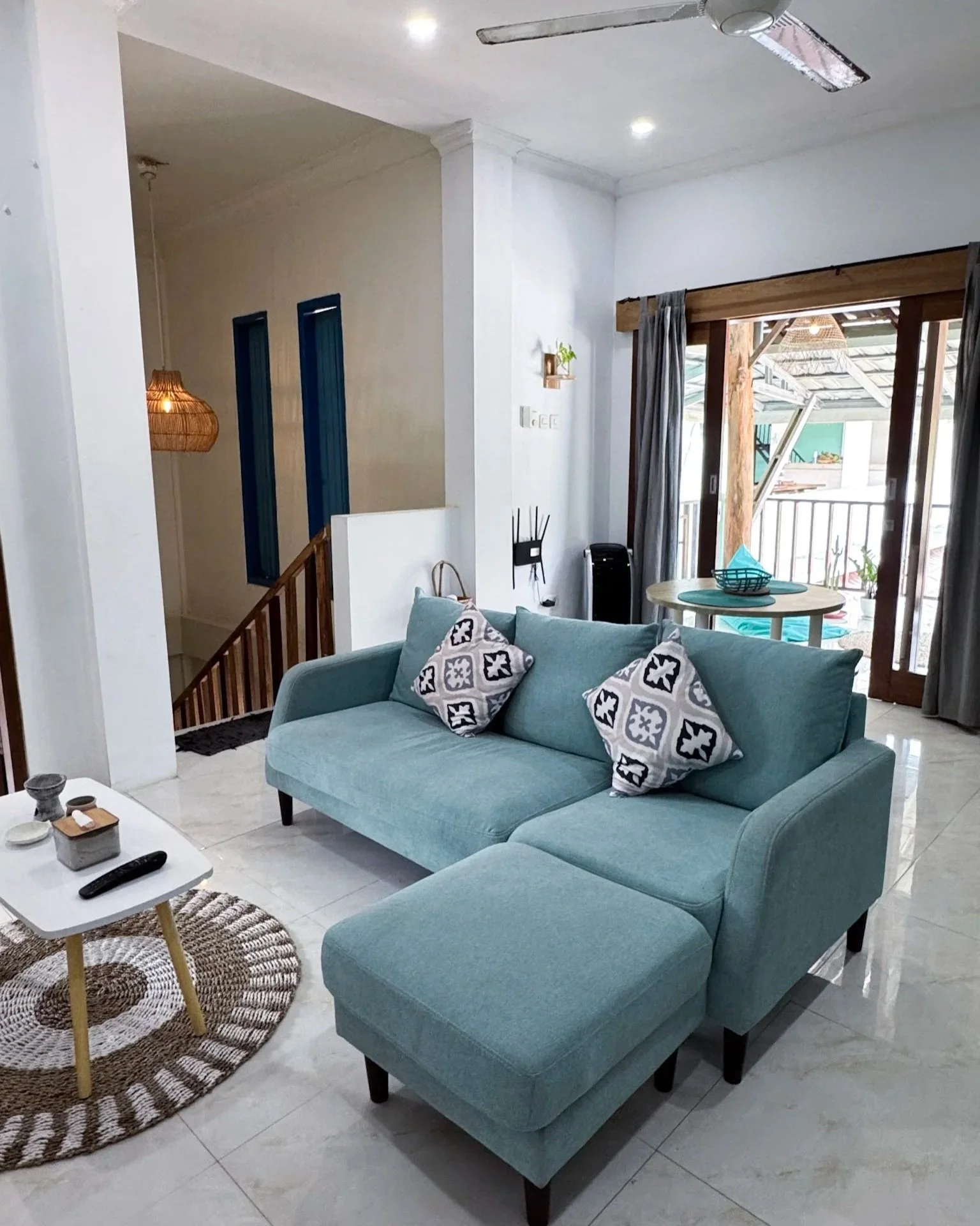 Living room with light blue sectional sofa with patterned throw pillows, round woven rug, white coffee table with remote and decor, sliding glass doors to outdoor balcony, ceiling fan, and modern decor.