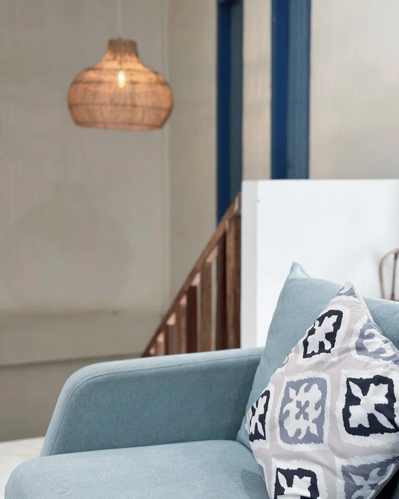 Close-up of a light blue sofa with a patterned pillow in a living room setting. In the background, there is a wicker pendant lamp hanging from the ceiling and a stair railing with wooden elements.