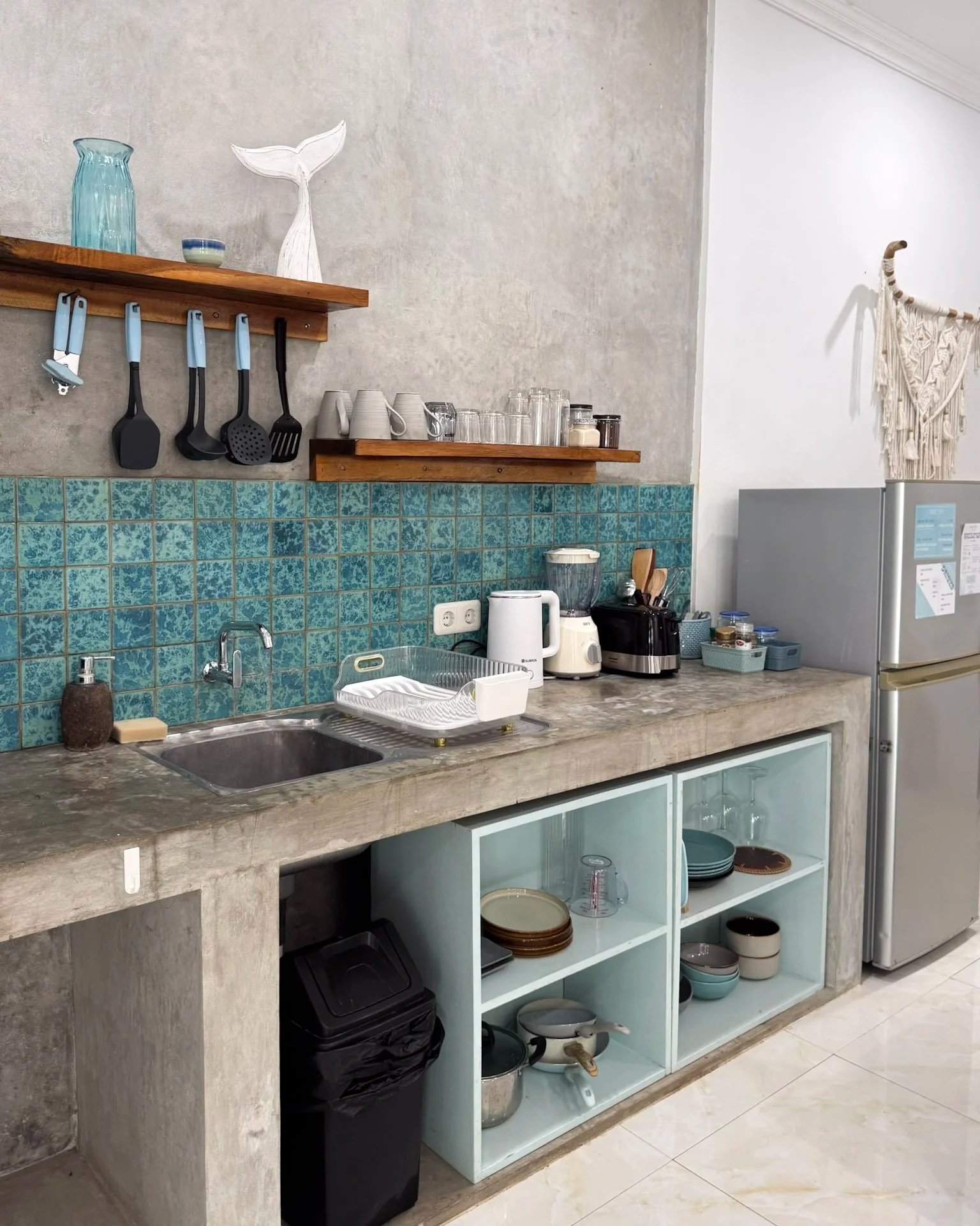 A kitchen with a concrete countertop, light blue open shelving with plates and bowls, a sink with a dish rack, and a bar area with mugs, glasses, and kitchen utensils on wooden shelves, a refrigerator with papers attached, and a wall decoration made 