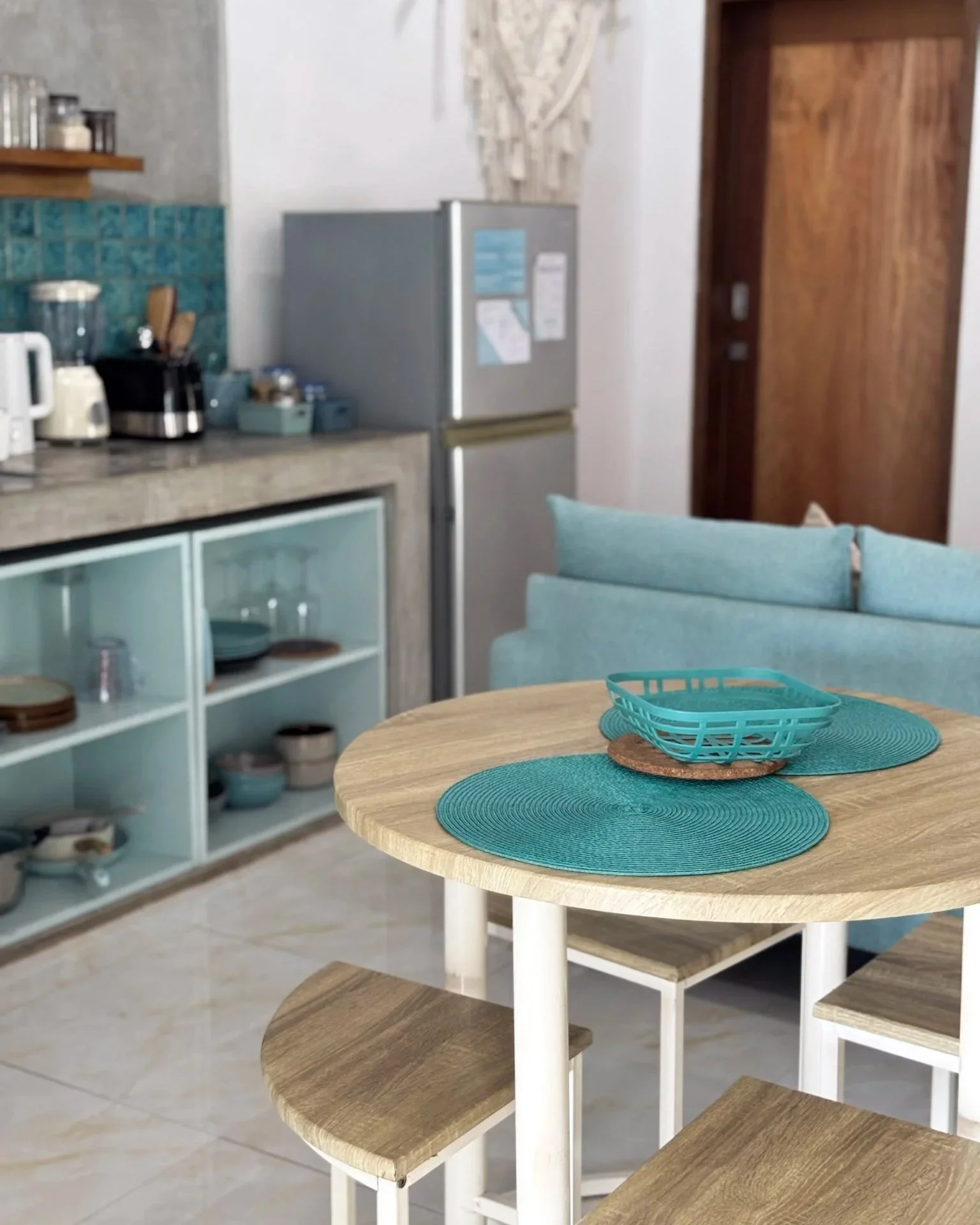 A cozy kitchen and dining area with a light wood round table, teal placemats, a teal basket, and matching teal seating. In the background, there is a partial view of a kitchen counter with appliances, an open shelving unit with dishes, a refrigerator