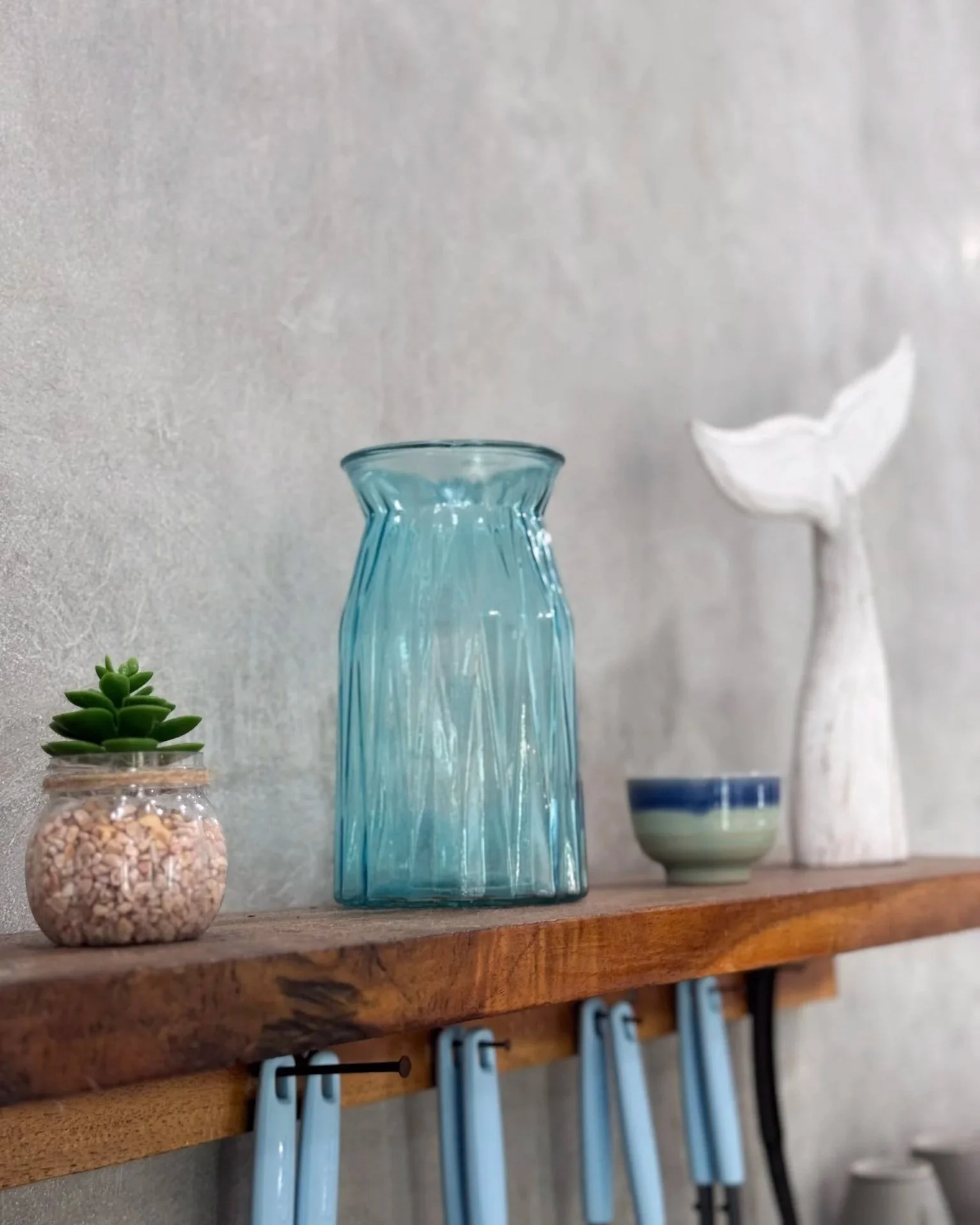Decorative objects on a wooden shelf including a small succulent in a glass jar, a tall blue glass vase, a small bowl with blue and green stripes, and a white ceramic whale sculpture.