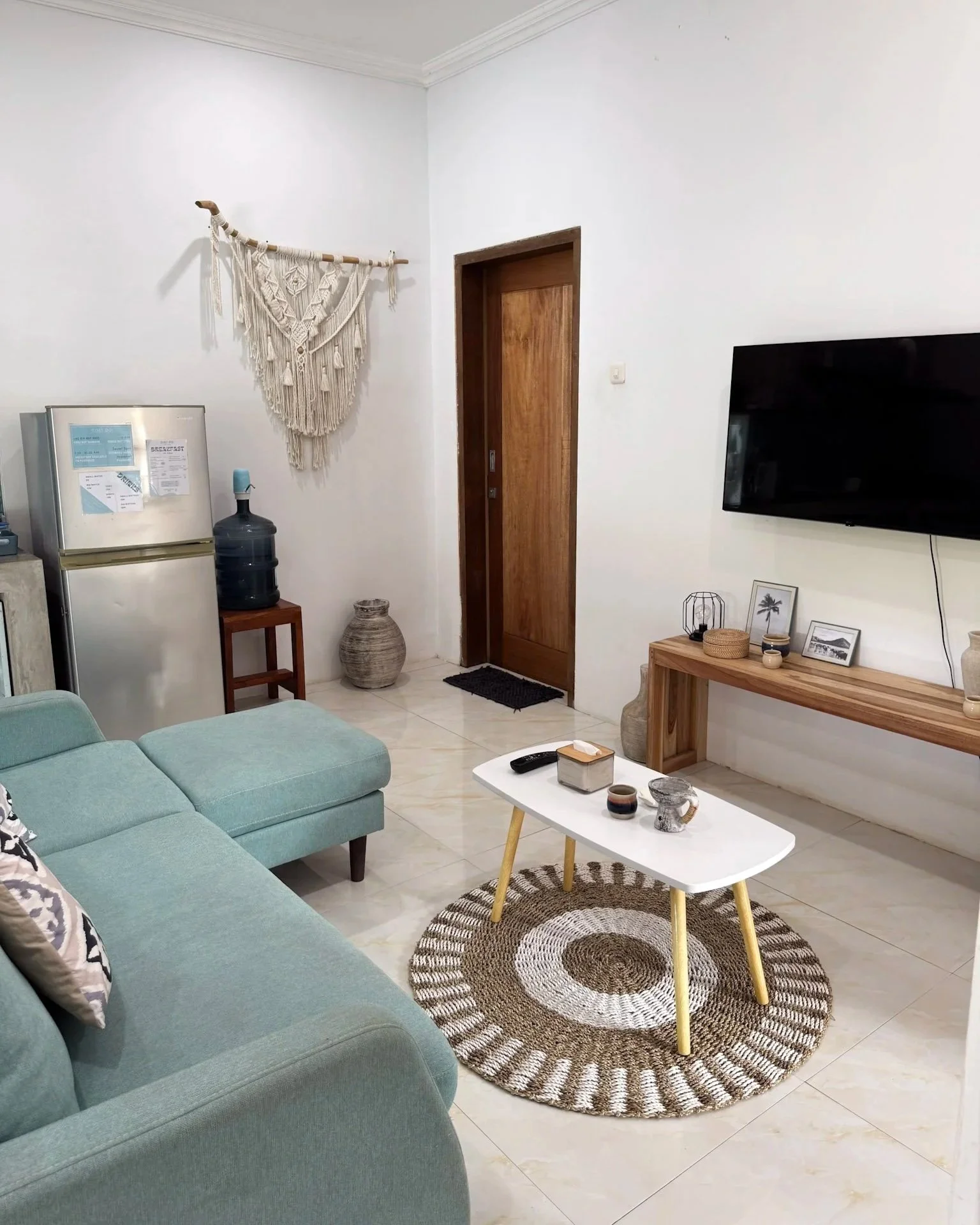 Living room with a teal sectional sofa, white coffee table on a circular woven rug, wall-mounted flat-screen TV, wooden console, and decorative items including framed pictures and vases. A door, a water dispenser, and a macrame wall hanging are also 