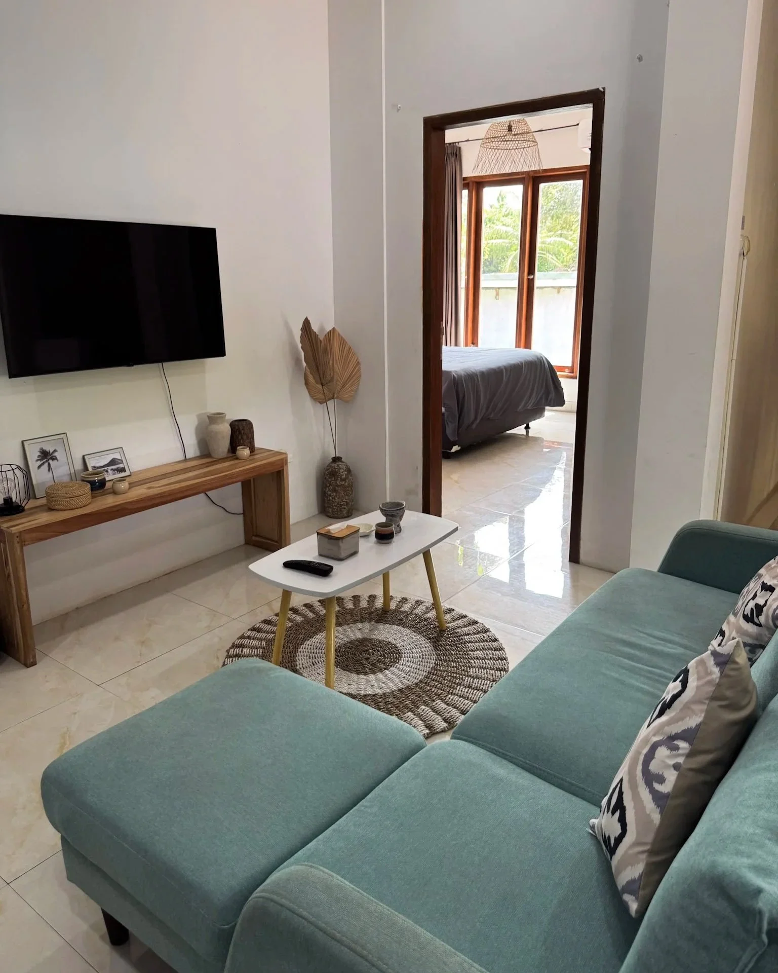 Living room with a turquoise sofa, a patterned pillow, a small white coffee table with a remote and decorative items, a wooden console with framed photo and decor, wall-mounted TV, and a view through a doorway to a bedroom with a bed and large window