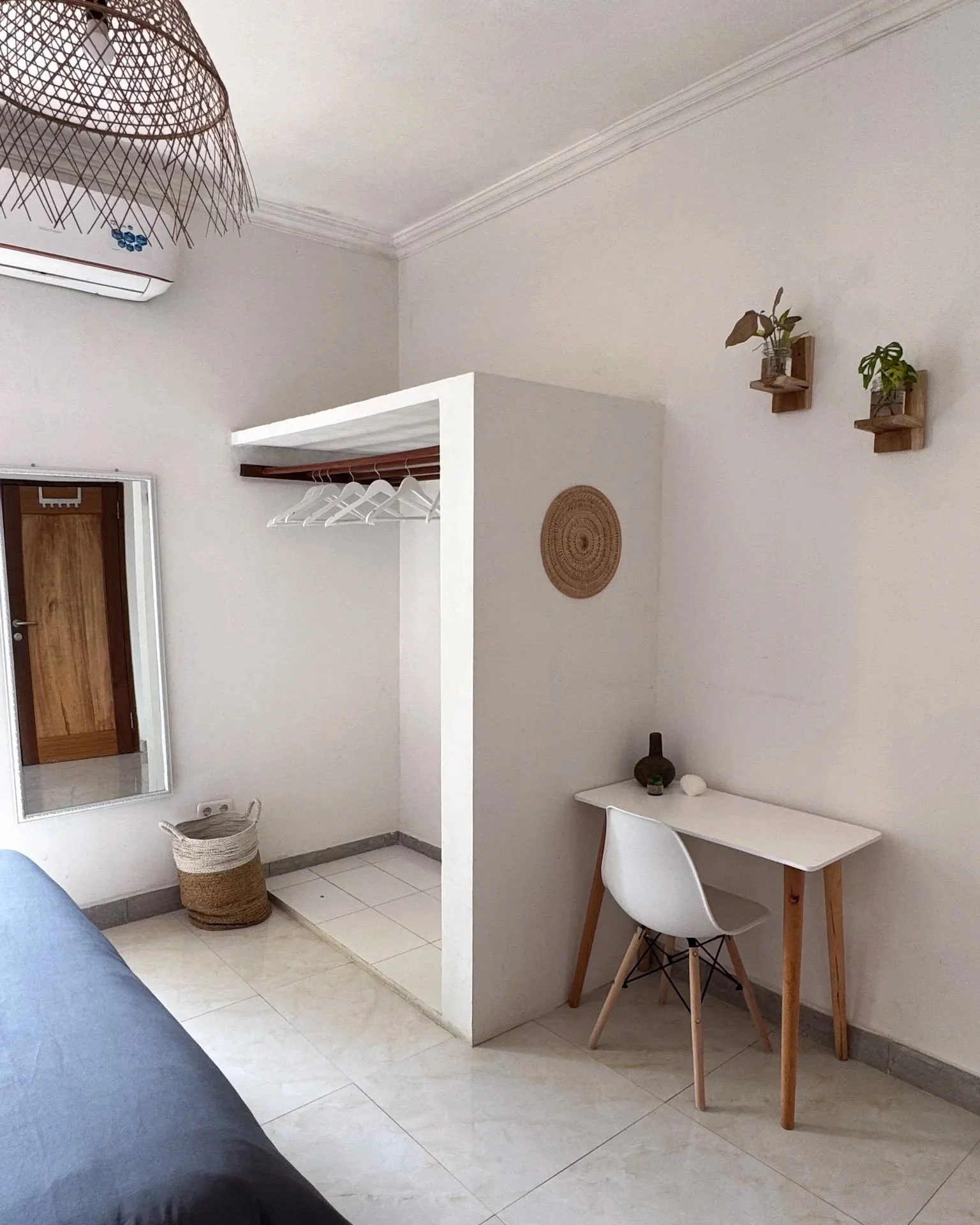 Minimalist bedroom corner with an open closet, a white desk with wooden legs, a white chair, decorative wall shelves with plants, and beige tiled flooring.