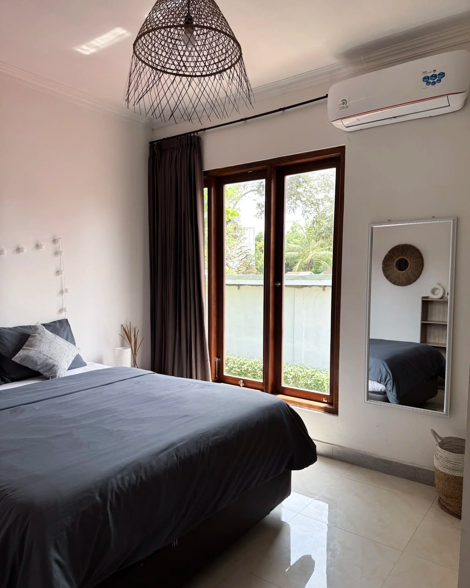 A bedroom with a bed, dark gray bedding, and pillows, next to a large window with wooden frames and dark curtains, a wall-mounted air conditioner, a mirror on the wall, and a wicker basket on the floor.
