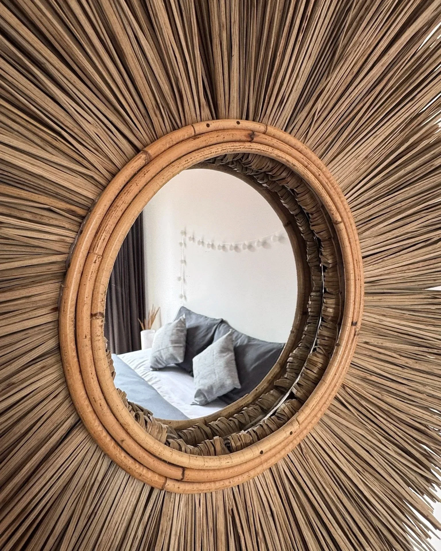 Decorative mirror with a woven rattan frame reflects a bedroom with pillows and string lights.