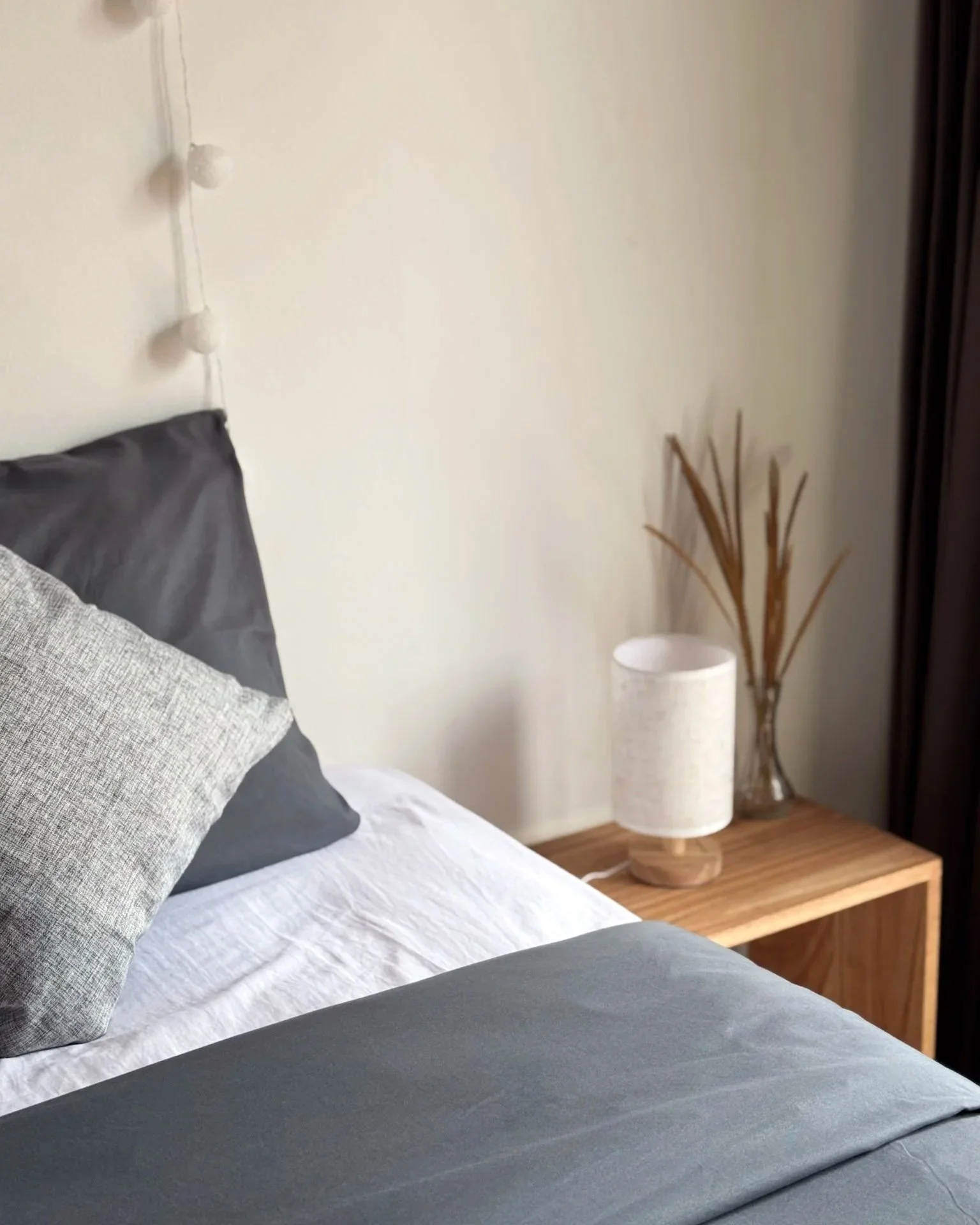 A neatly made bed with gray and white pillows, a white sheet, and a gray blanket. To the side, there is a wooden nightstand with a white table lamp and a glass vase with dried tall grasses. A string of white spherical lights hangs on the wall above t