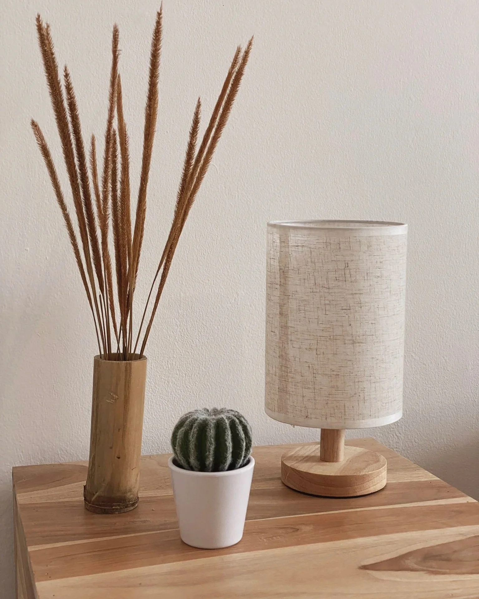 A wooden table with a tall glass vase holding dried grass, a small potted cactus, and a wooden table lamp with a fabric shade.