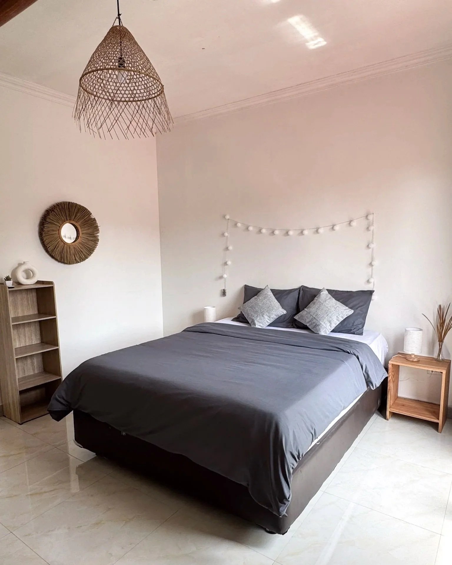 A neatly made bed with gray bedding and pillows, a small wooden nightstand with a lamp and decorative plant, a wooden shelf with decor, a circular mirror, and a rattan pendant light hanging from the ceiling in a minimalist bedroom.