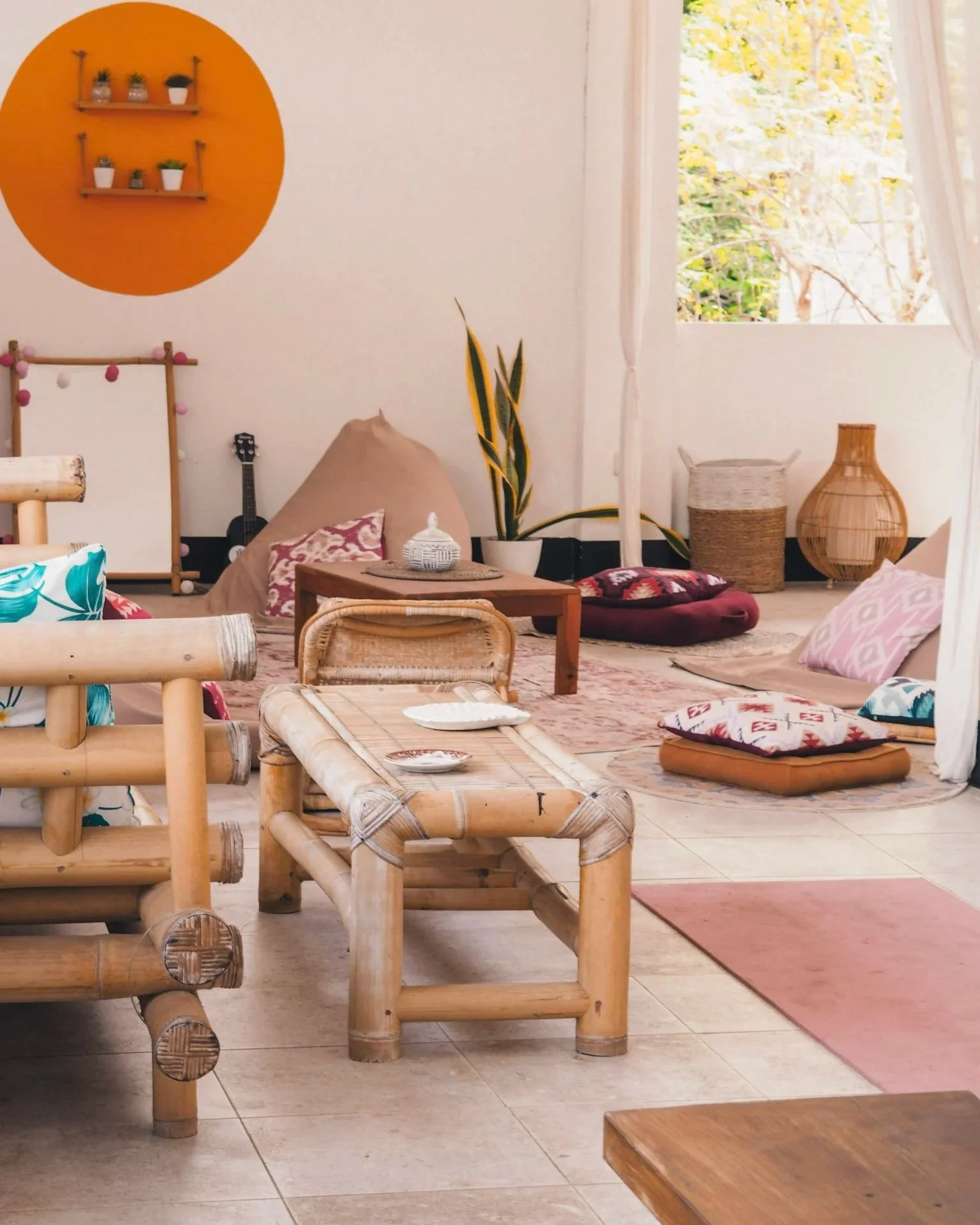 Cozy indoor space with tropical bamboo furniture, pillows, plants, and a window showing greenery outside.