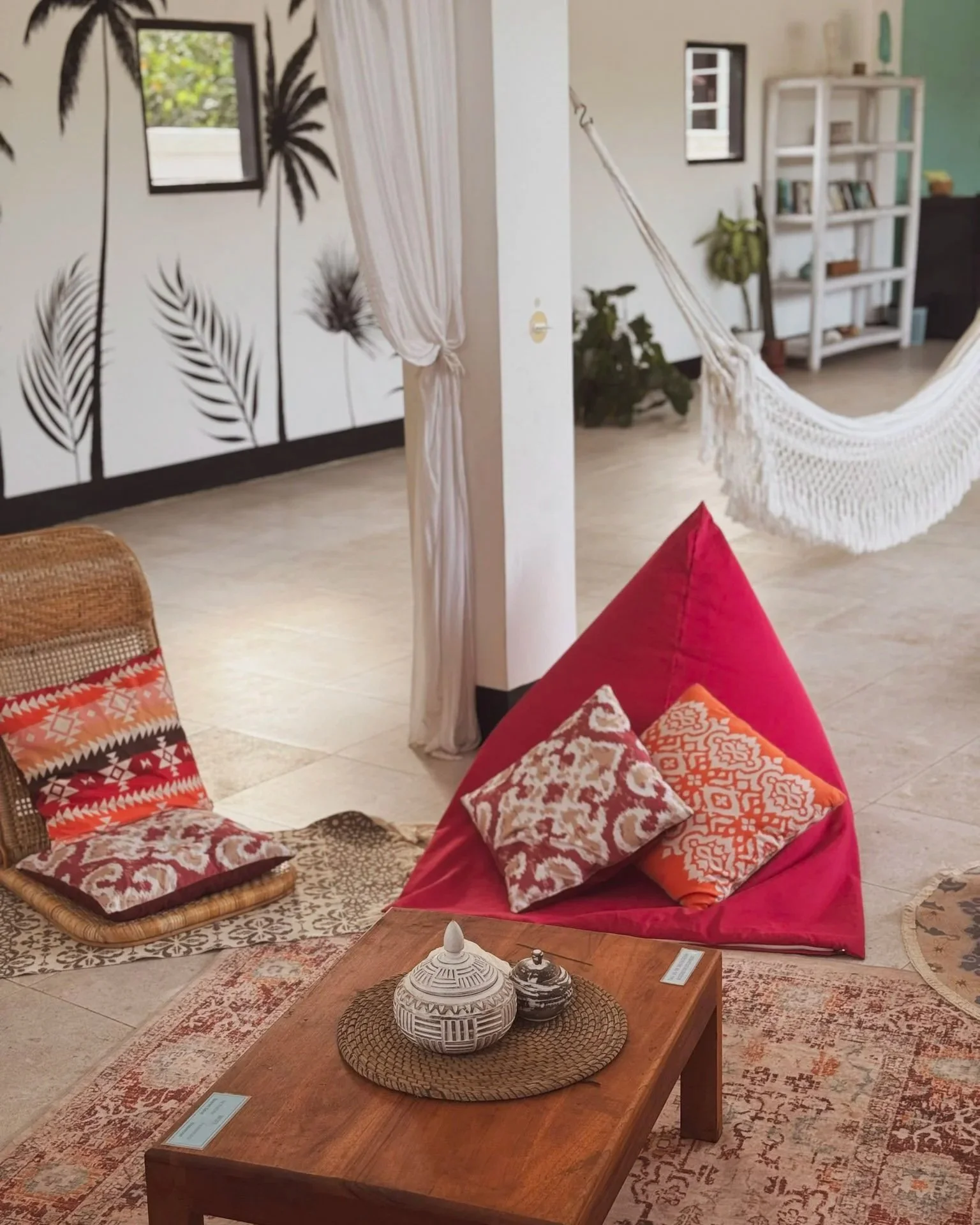 A cozy living space decorated with bohemian-inspired cushions and a hammock, with a wall featuring black palm tree silhouettes and a small mirrored window.