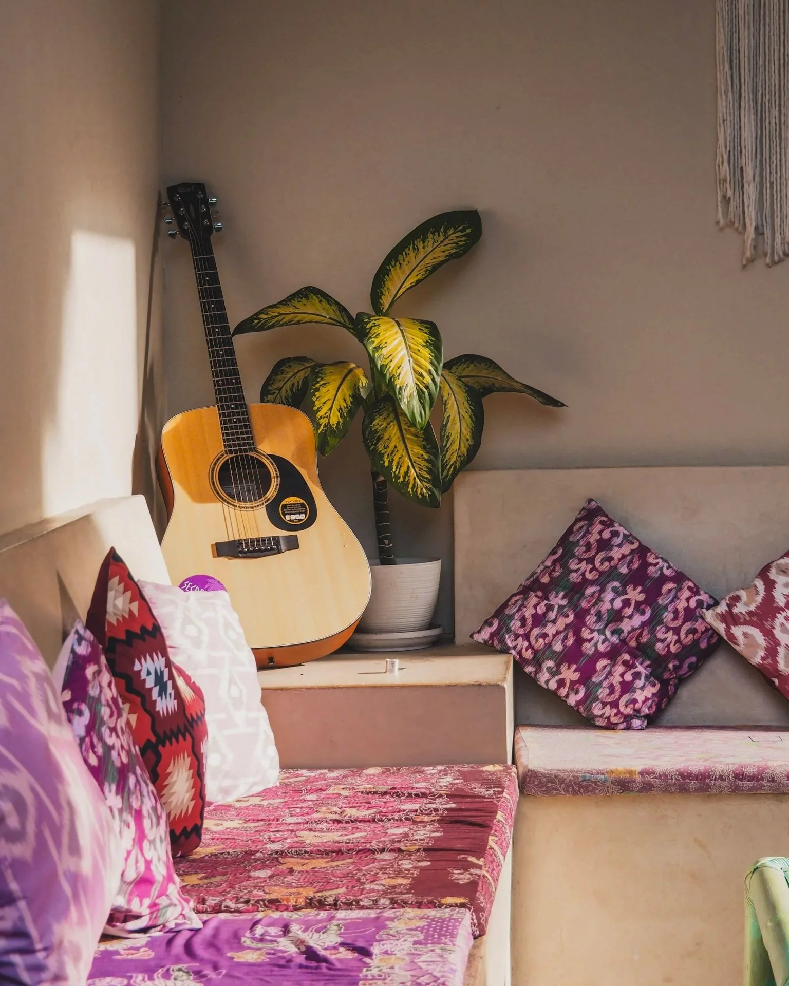 A room corner with a beige sofa, colorful patterned pillows, an acoustic guitar leaning against a wall, a potted plant with large green and yellow leaves, and a wall hanging on the right.
