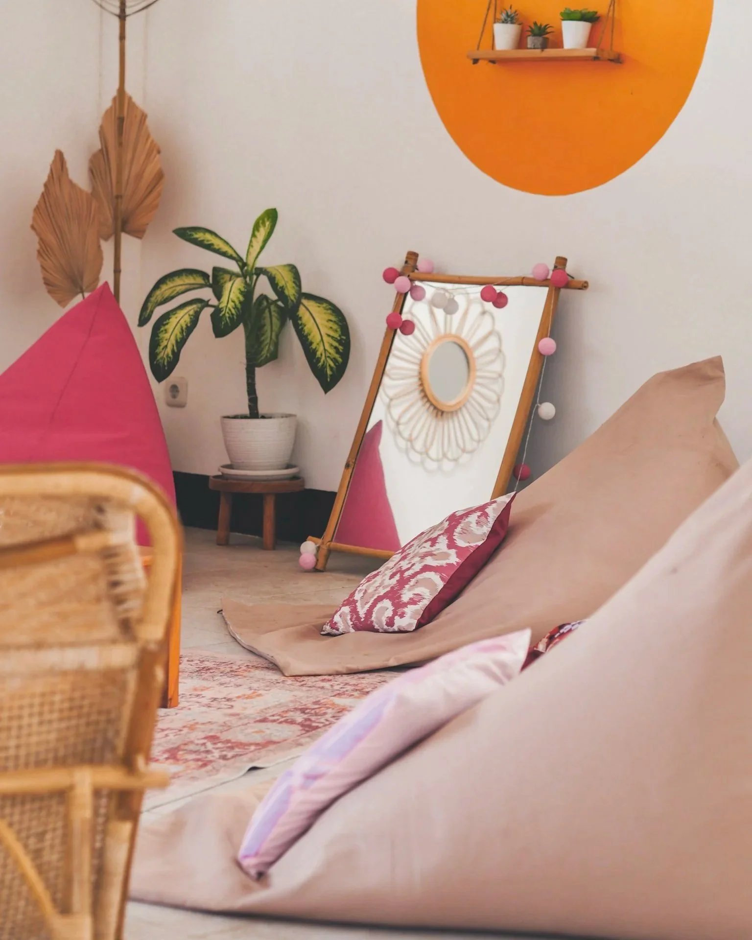 Cozy living room corner with cushions on the floor, decorative pillows, a mirror with pom-pom garland, a potted plant, woven wall art, and wall shelves with small plants