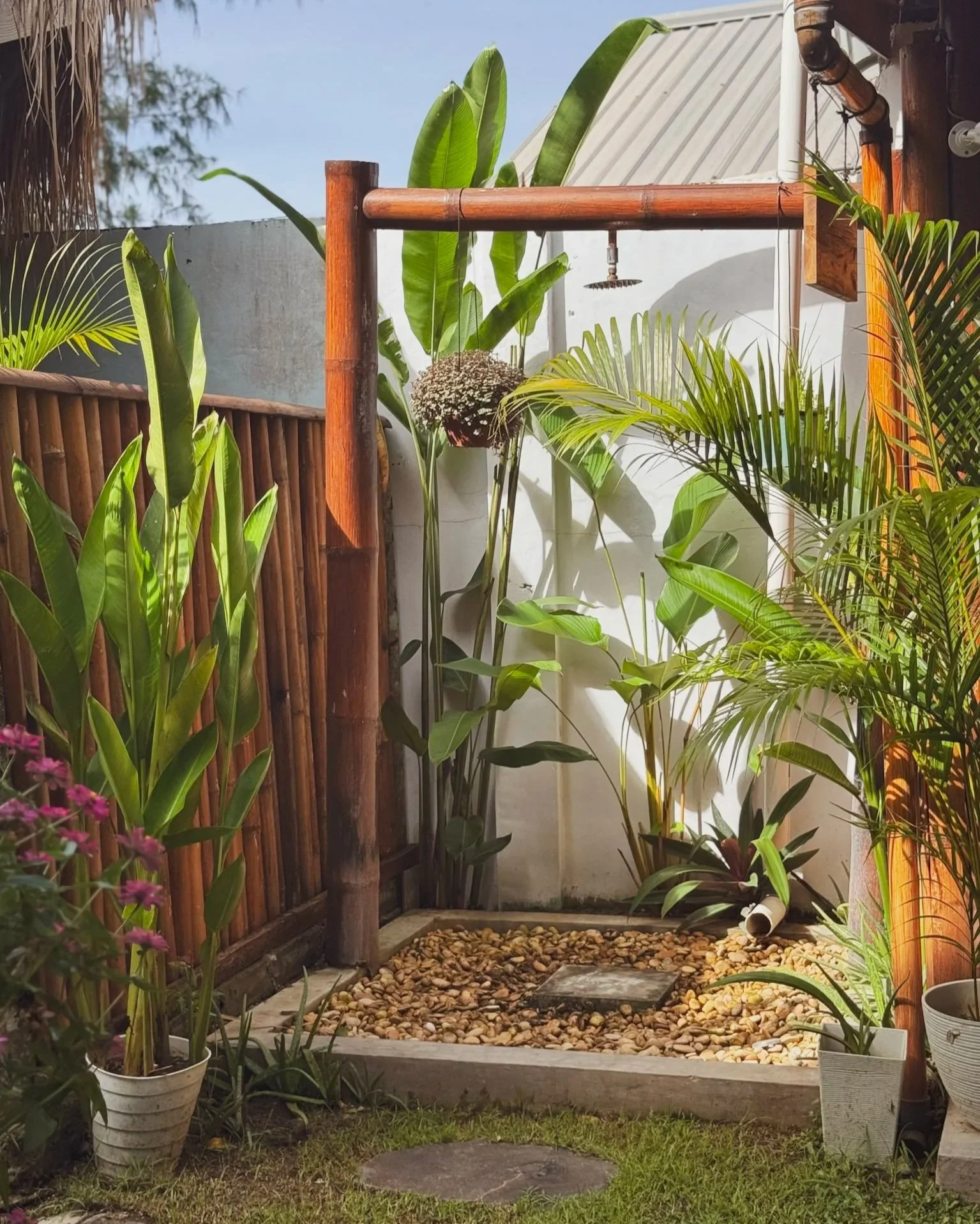 Small outdoor shower with a wooden support beam, surrounded by tropical plants and rocks, with a white pipe on the ground and a drain pipe in the corner.