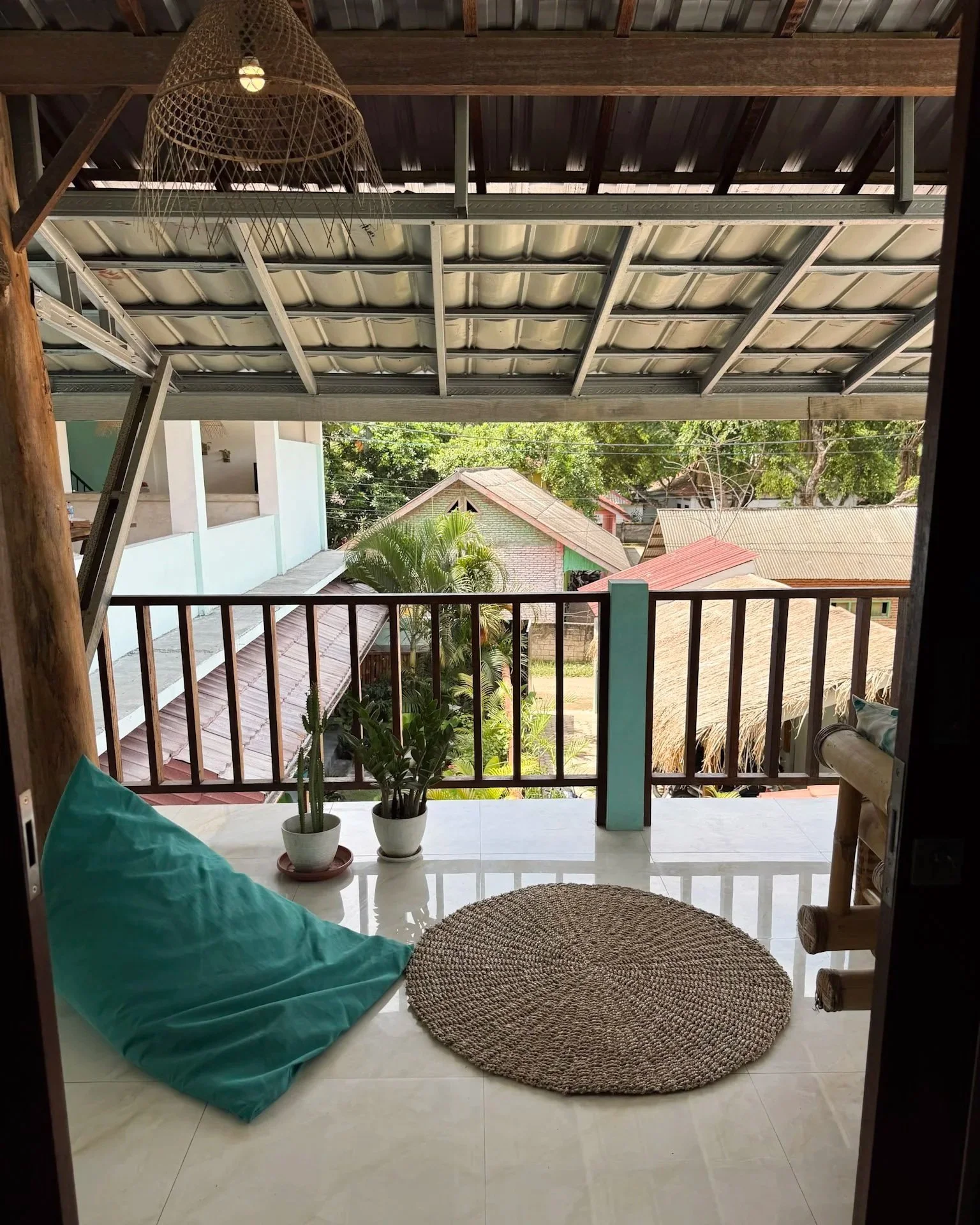 Balcony with a teal pillow on the floor, two potted plants, a round woven rug, and a view of trees and rooftops outside.