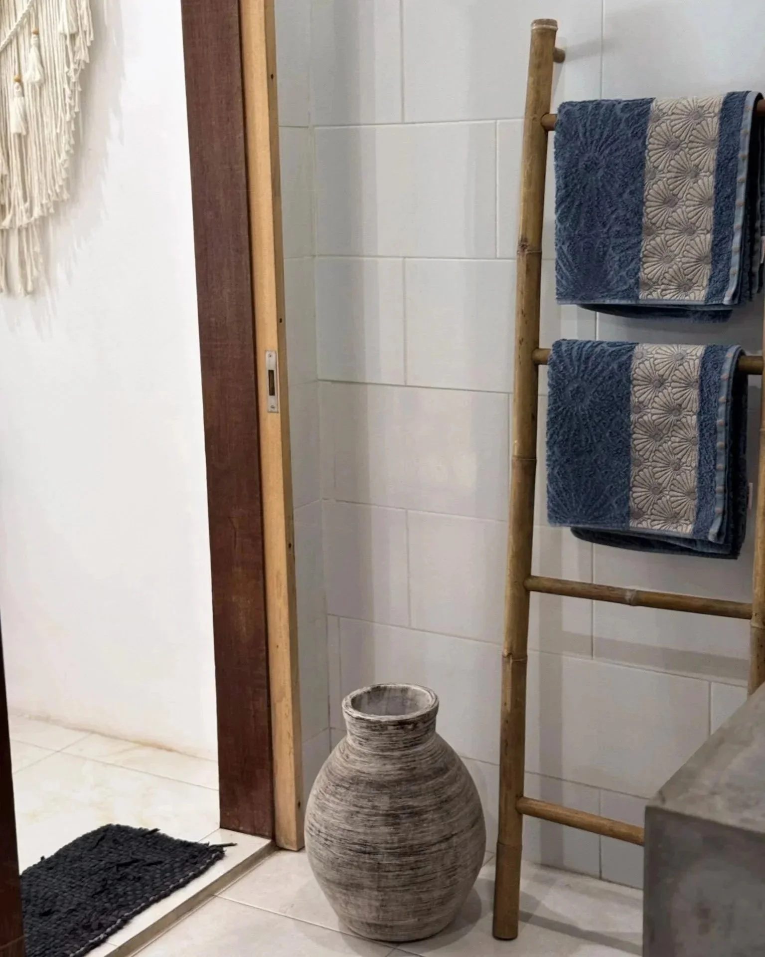 Interior view of a bathroom with a bamboo towel ladder holding two blue towels, a large rustic vase on the floor, and part of a shower area with a sliding wooden door.