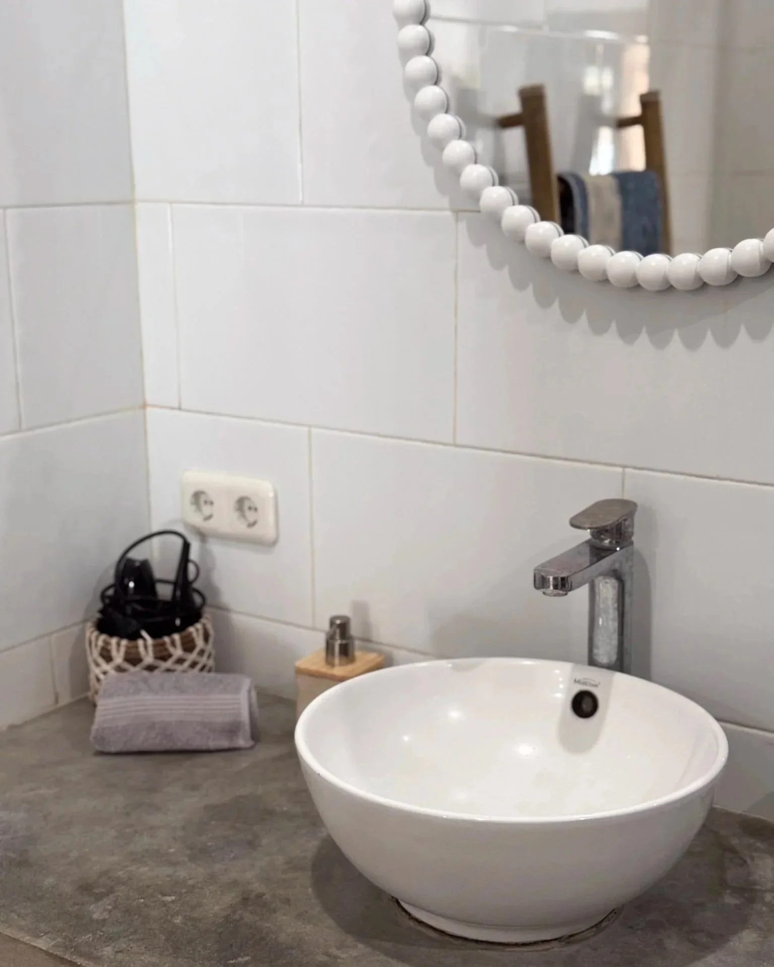 Bathroom countertop with a round white vessel sink, a chrome faucet, a soap dispenser, a woven basket holding black cords, a gray towel, and a wall plug, with a mirror and wooden towel racks reflected in the mirror.