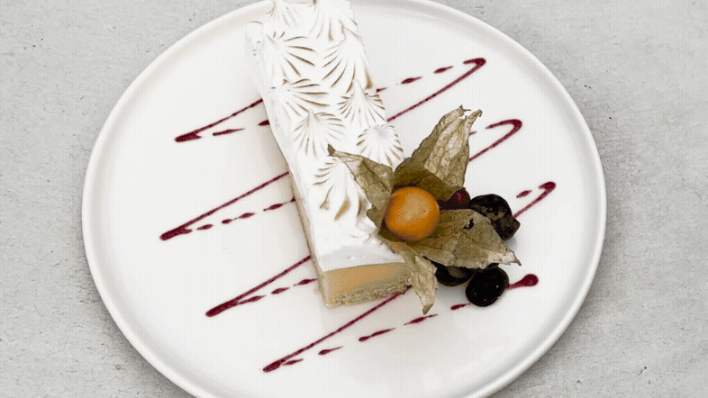 Porción de pastel de queso con decoraciones de frutos rojos y hoja de physalis sobre plato blanco con salsa de frutos rojos.