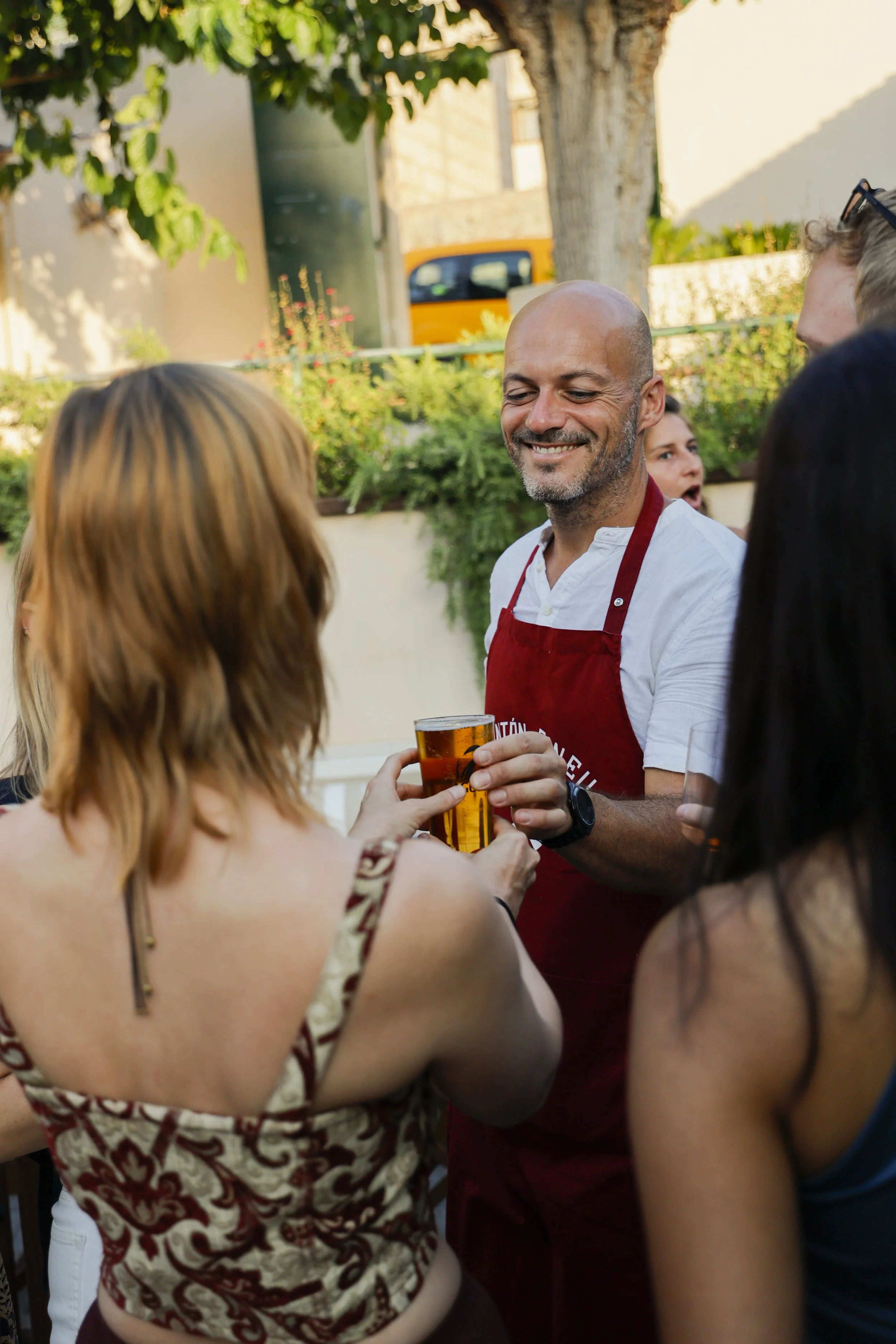 Escena de personas en una celebración al aire libre, con un hombre sirviendo y entregando una bebida a una mujer, en un entorno con árboles y plantas.