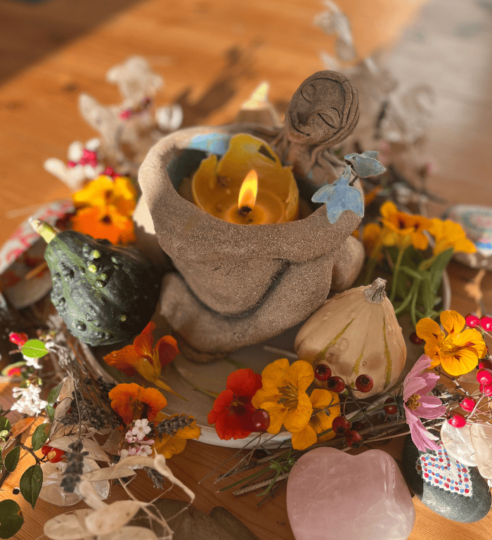 The holding woman candle holding a candle in her arms in the centre of a plate of flowers and veg, surrounded by crystals - at a circle hosted by Doula Harriet Symon.