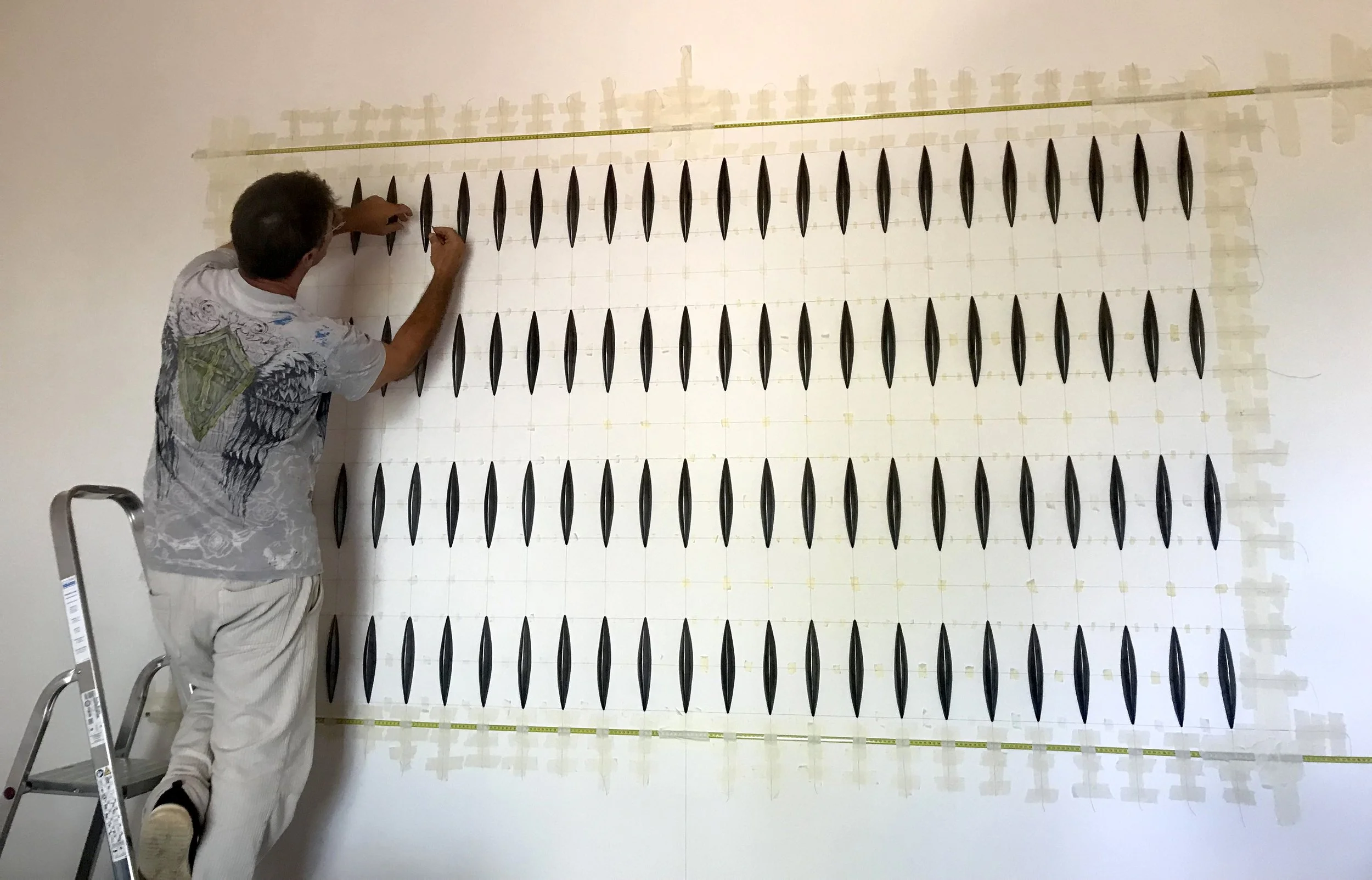 Man standing on a ladder, installing or adjusting a grid of black oval objects on a wall, possibly part of an art installation or scientific display.