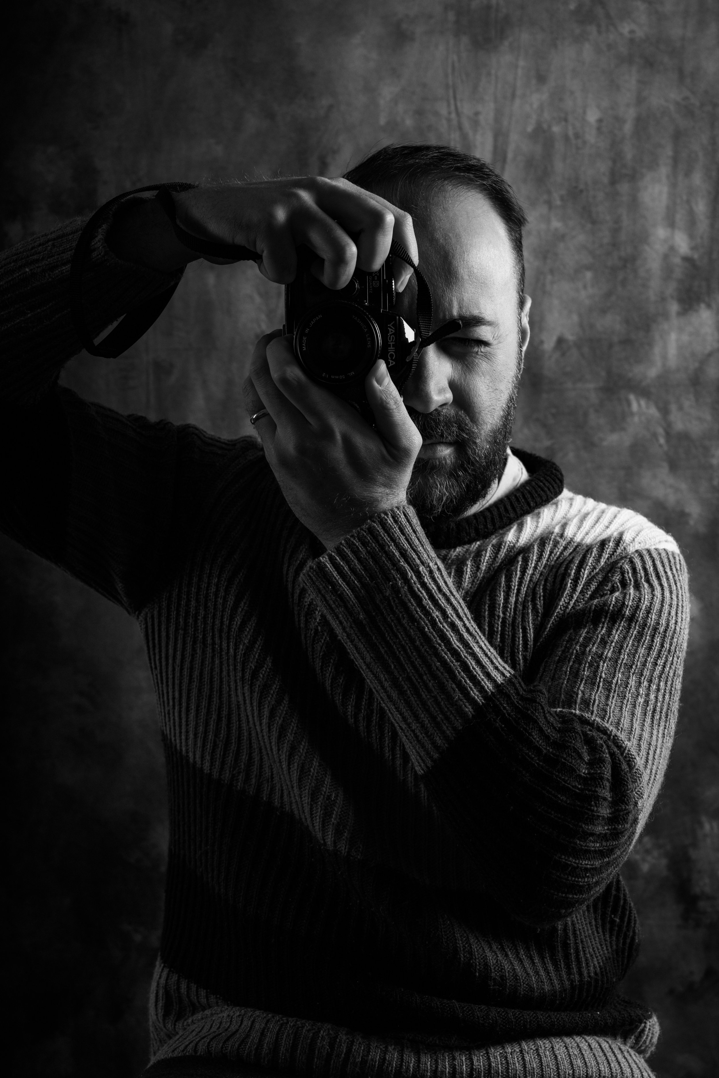 Un uomo con barba, che indossa un maglione a righe, si sta prendendo una foto riflessa in uno specchio, lui tiene una fotocamera davanti al volto. L'immagine è in bianco e nero e lo sfondo è semplice e scuro.