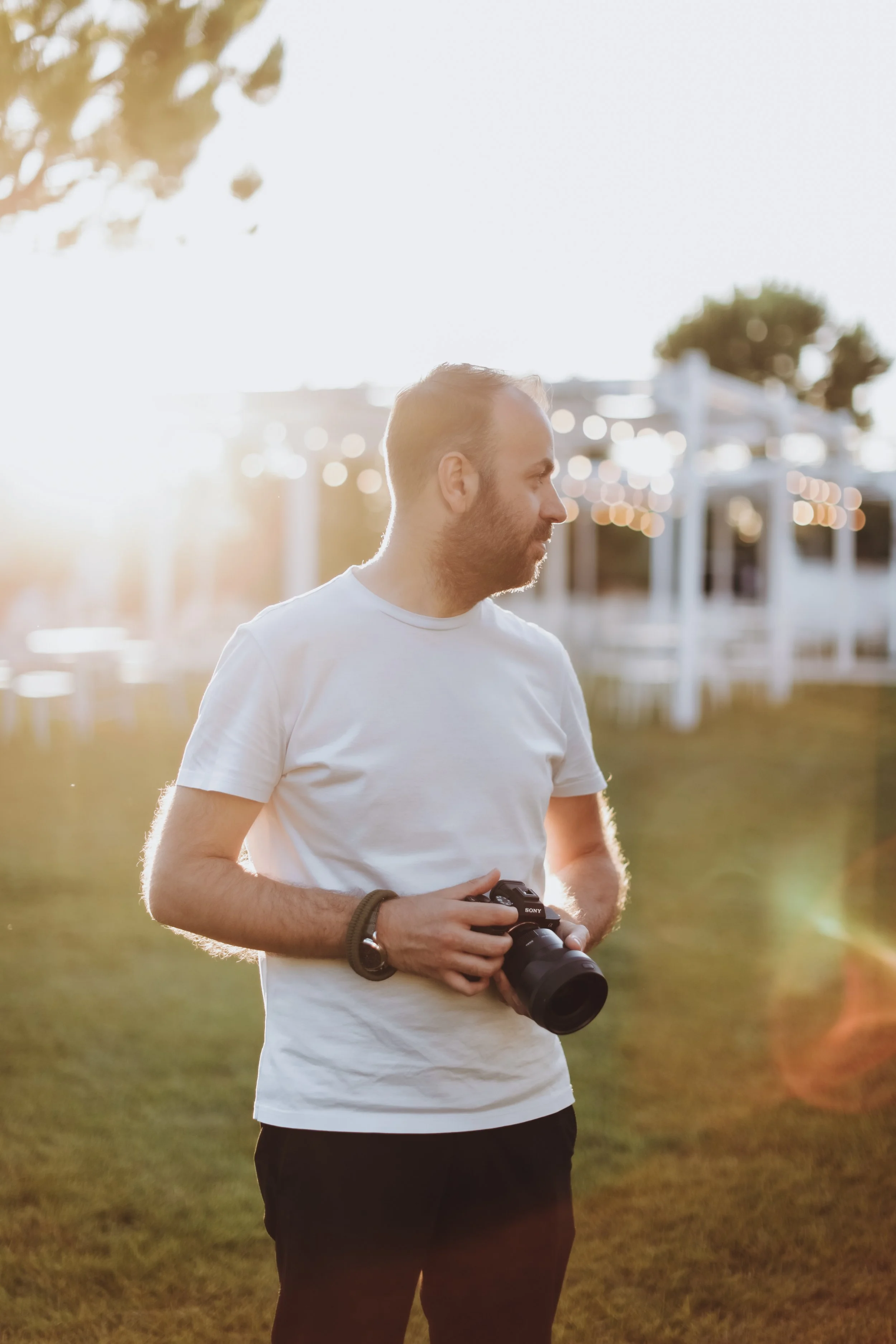Uomo con barba, maglietta bianca e pantaloni neri, che tiene una macchina fotografica Sony, si trova in un parco durante il tramonto.