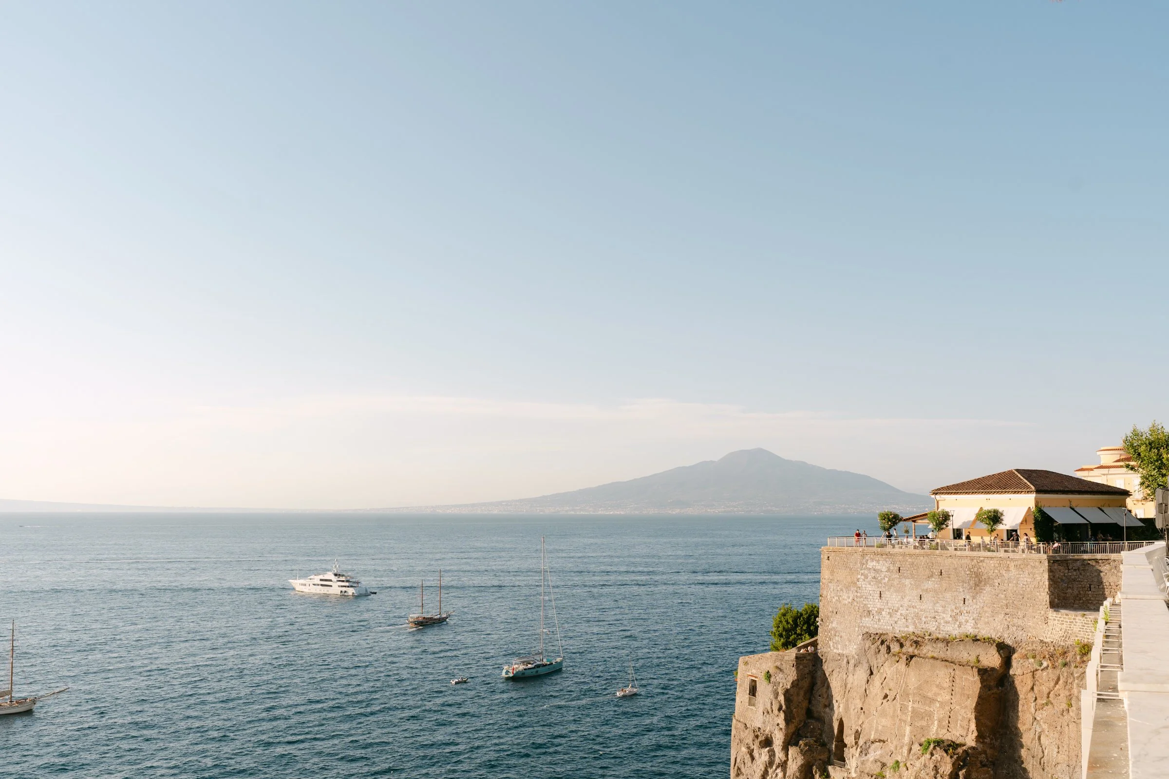 Vista panoramica del mare con alcune barche a vela e una grande nave, sullo sfondo un monte e un edificio con terrazza sul lungomare, alcuni alberi e persone che si godono la vista.
