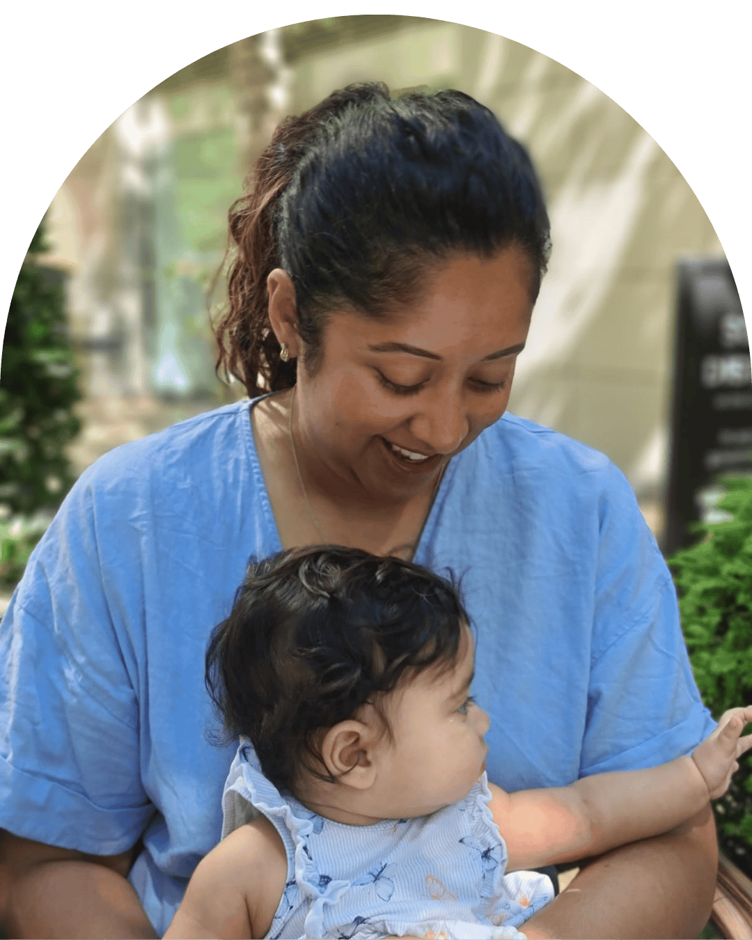 A woman in a blue shirt holding a young child with dark hair, both smiling and looking at each other indoors with plants around.