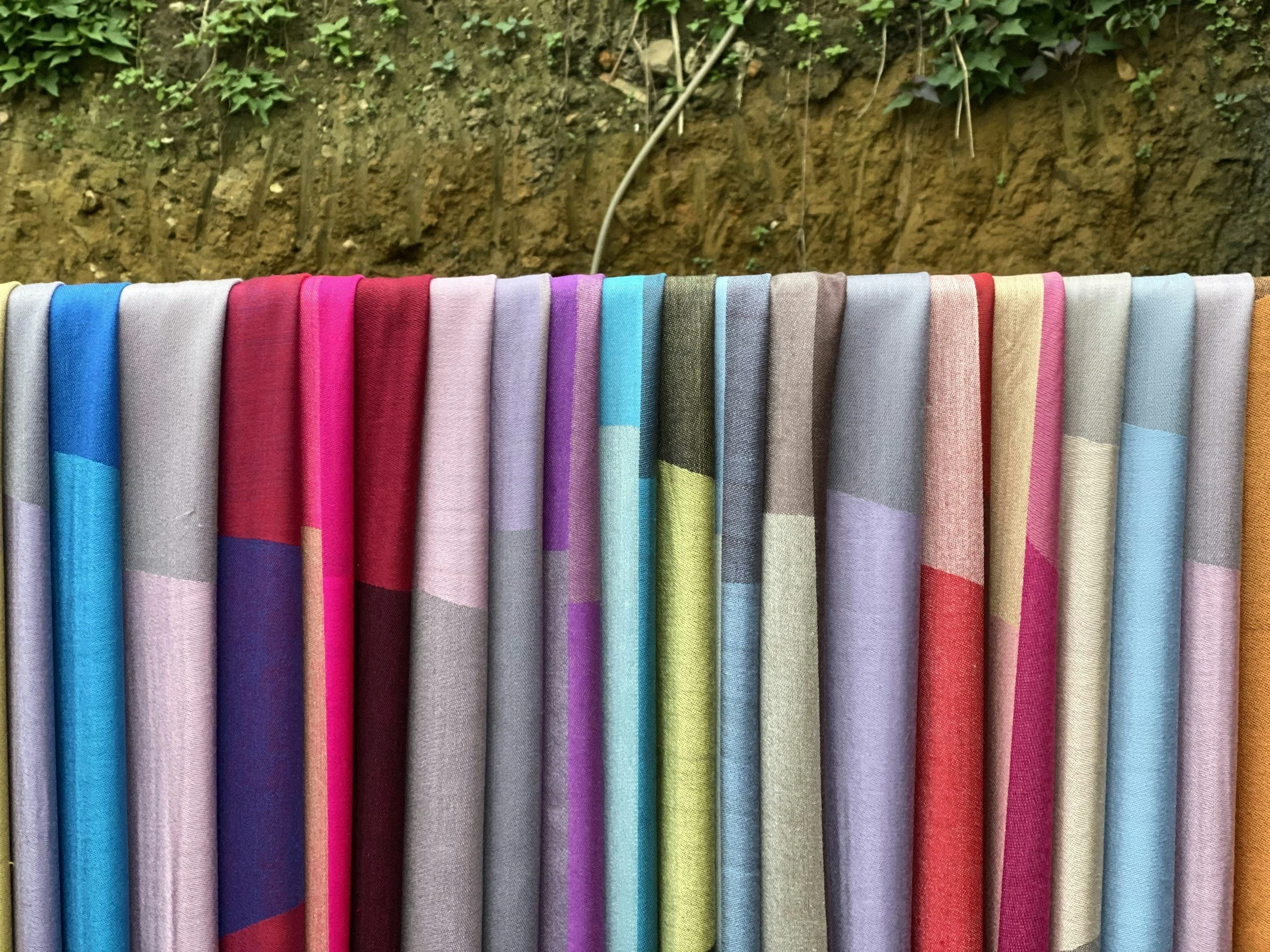 Colorful folded fabrics hanging outdoors with soil and plants in the background.