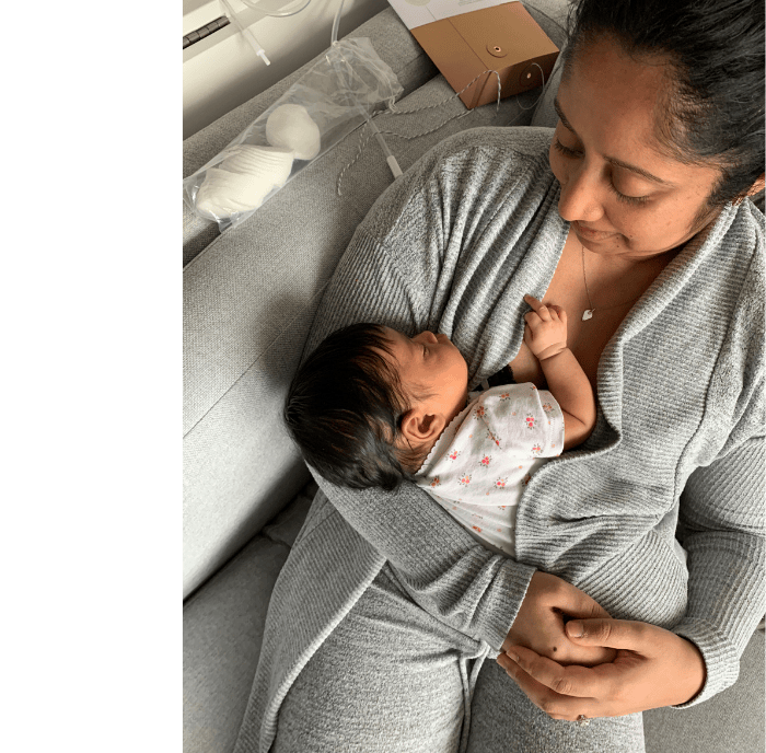 A woman is sitting on a gray couch with a baby lying on her chest. The woman is looking down at the baby, who is dressed in a white outfit with small prints.