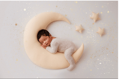 A baby sleeping on a crescent moon-shaped pillow with stars around, on a light background.